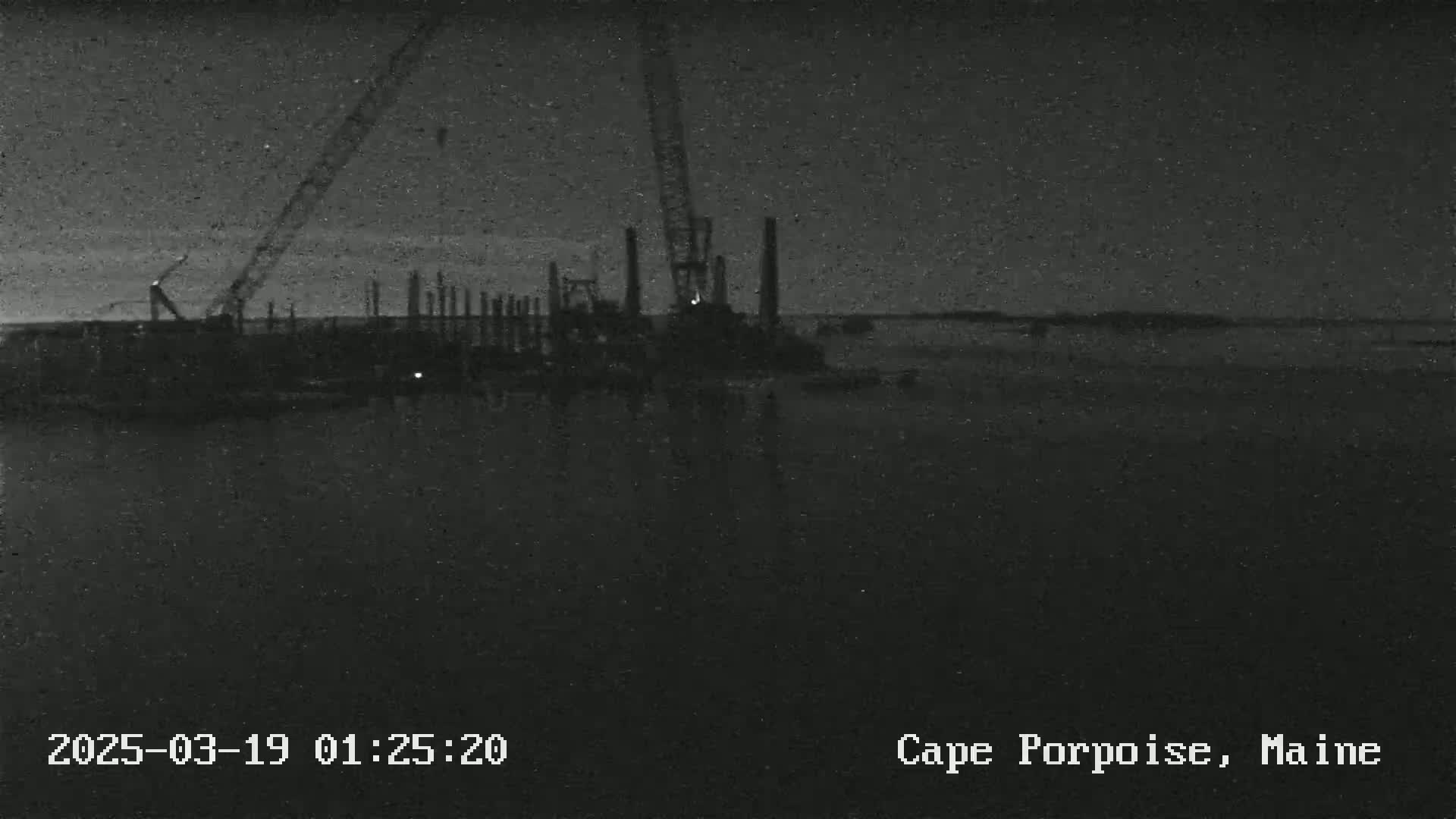 A nighttime, low-light view of construction equipment and pilings near a body of water.