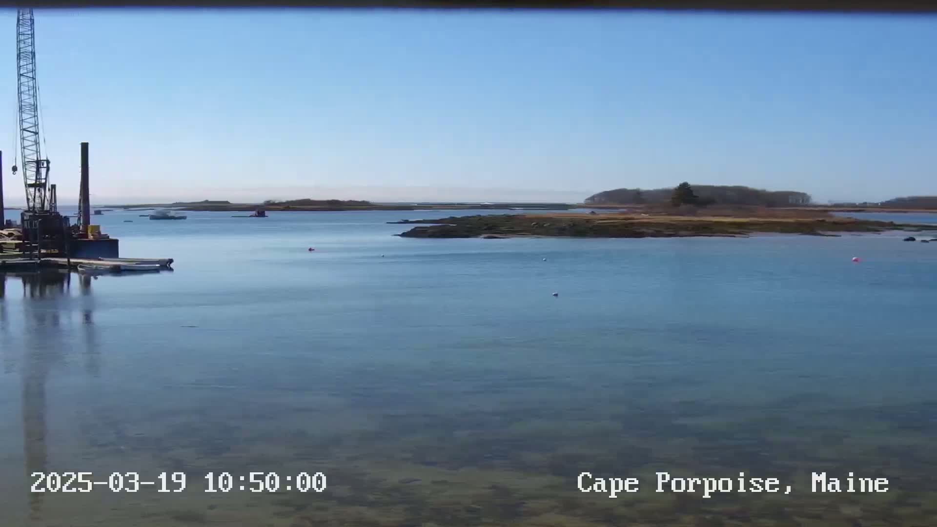 A calm, clear day shows a body of water with a crane and several boats near a shoreline, and small islands in the distance.