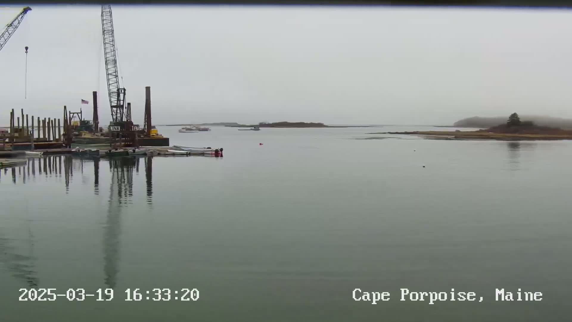 A foggy harbor scene shows construction cranes and several boats near a calm body of water with a distant shoreline.