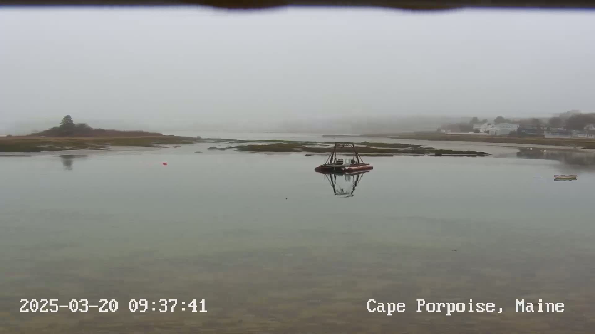 A calm body of water, shrouded in fog, shows a small dredging vessel and a few distant buildings on a foggy day.