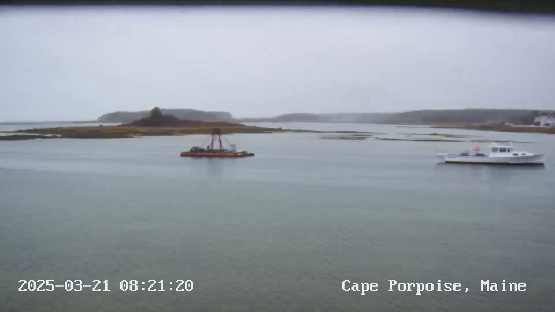 A calm, overcast day shows a small boat and a barge in a bay near a small, tree-covered island.