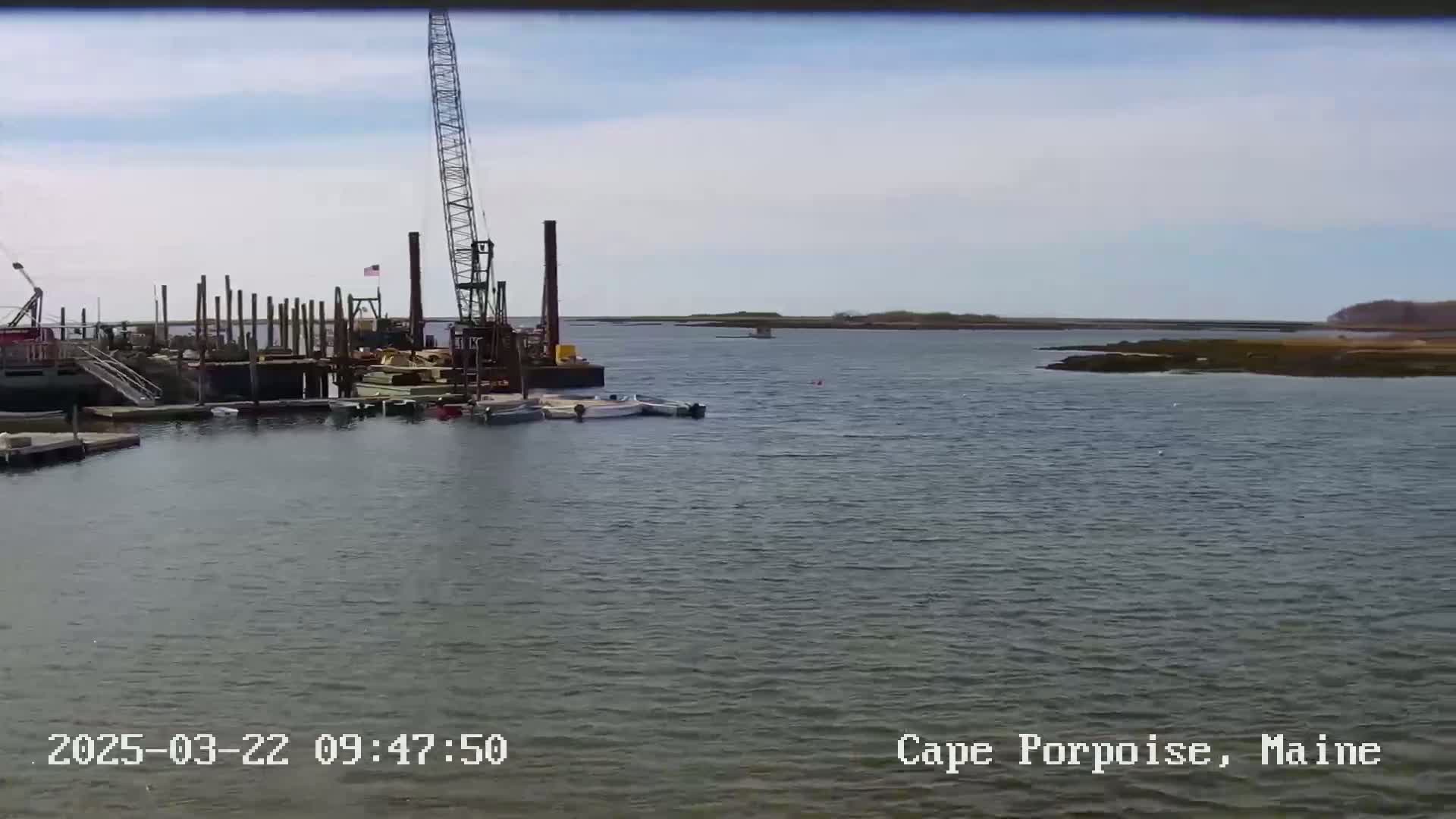 A large crane sits on a barge near a dock in a calm body of water under a mostly cloudy sky.