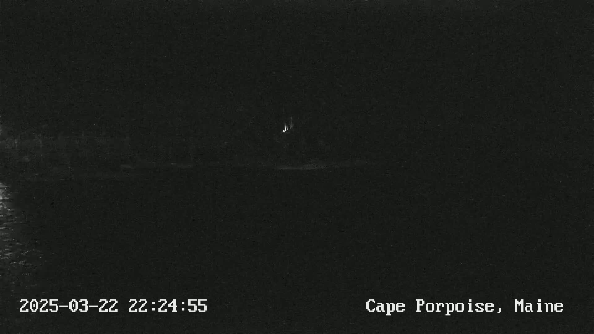 A dark, nighttime image shows a faint light source in the distance near a dark body of water.