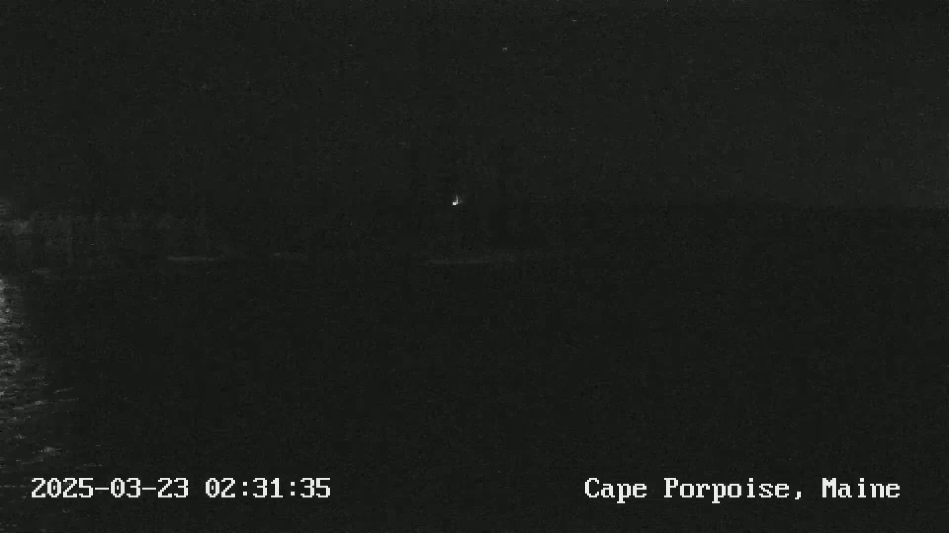 A dark, nighttime scene shows a faint light in the distance over dark water.