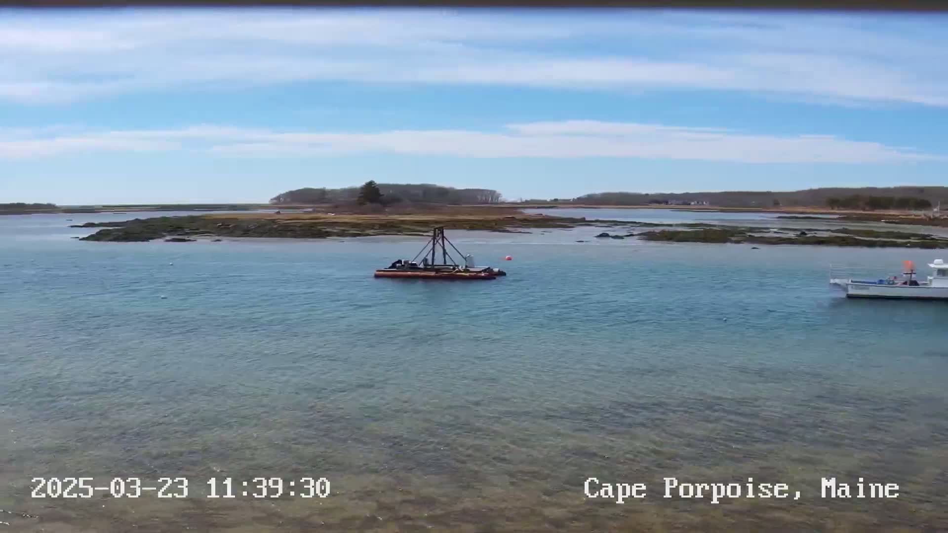 A small boat and a piece of work equipment are visible on calm, clear water near a rocky shoreline under a mostly sunny sky.