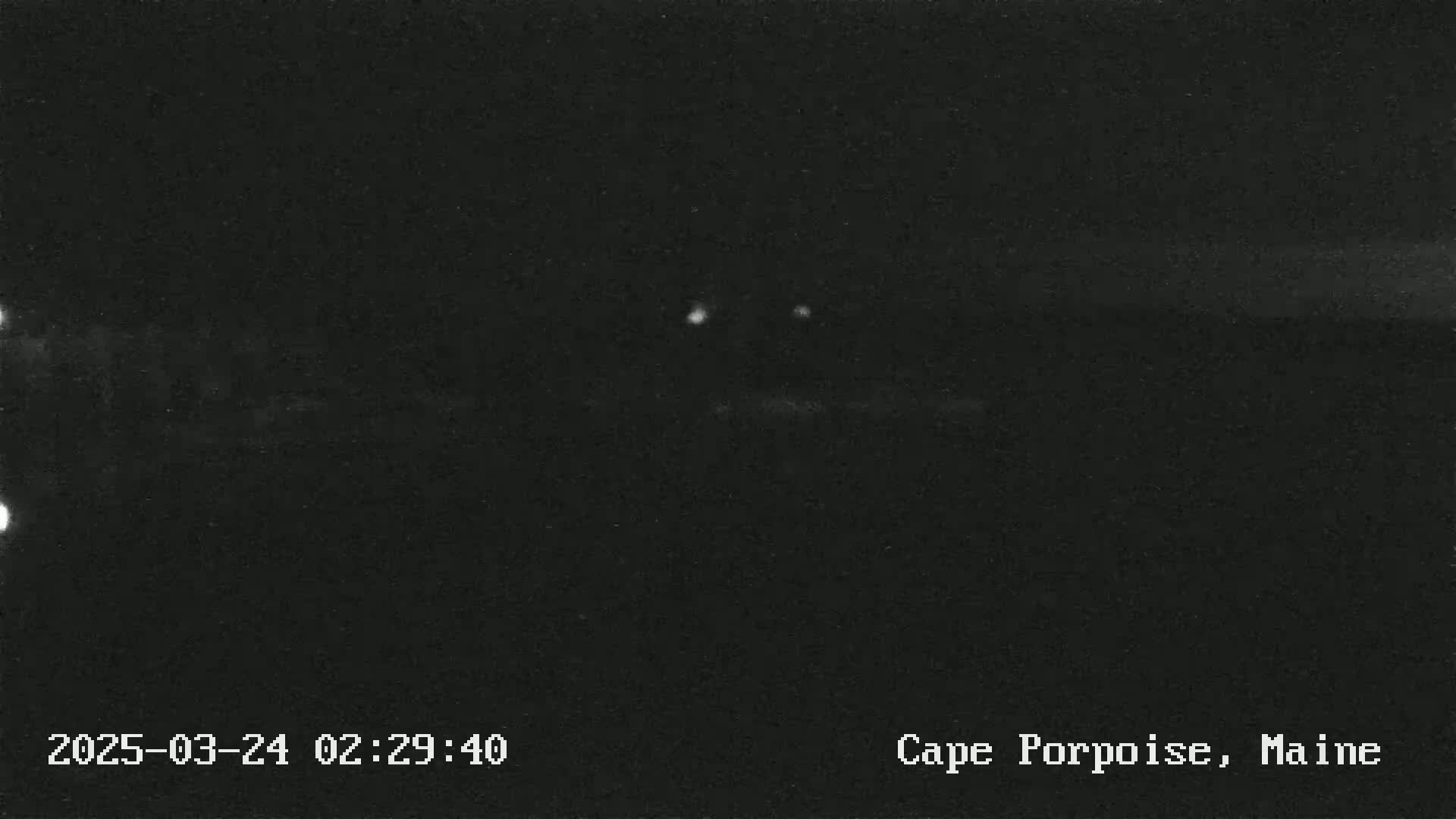 A nighttime, dark image shows a few faint lights in the distance under a clear sky.