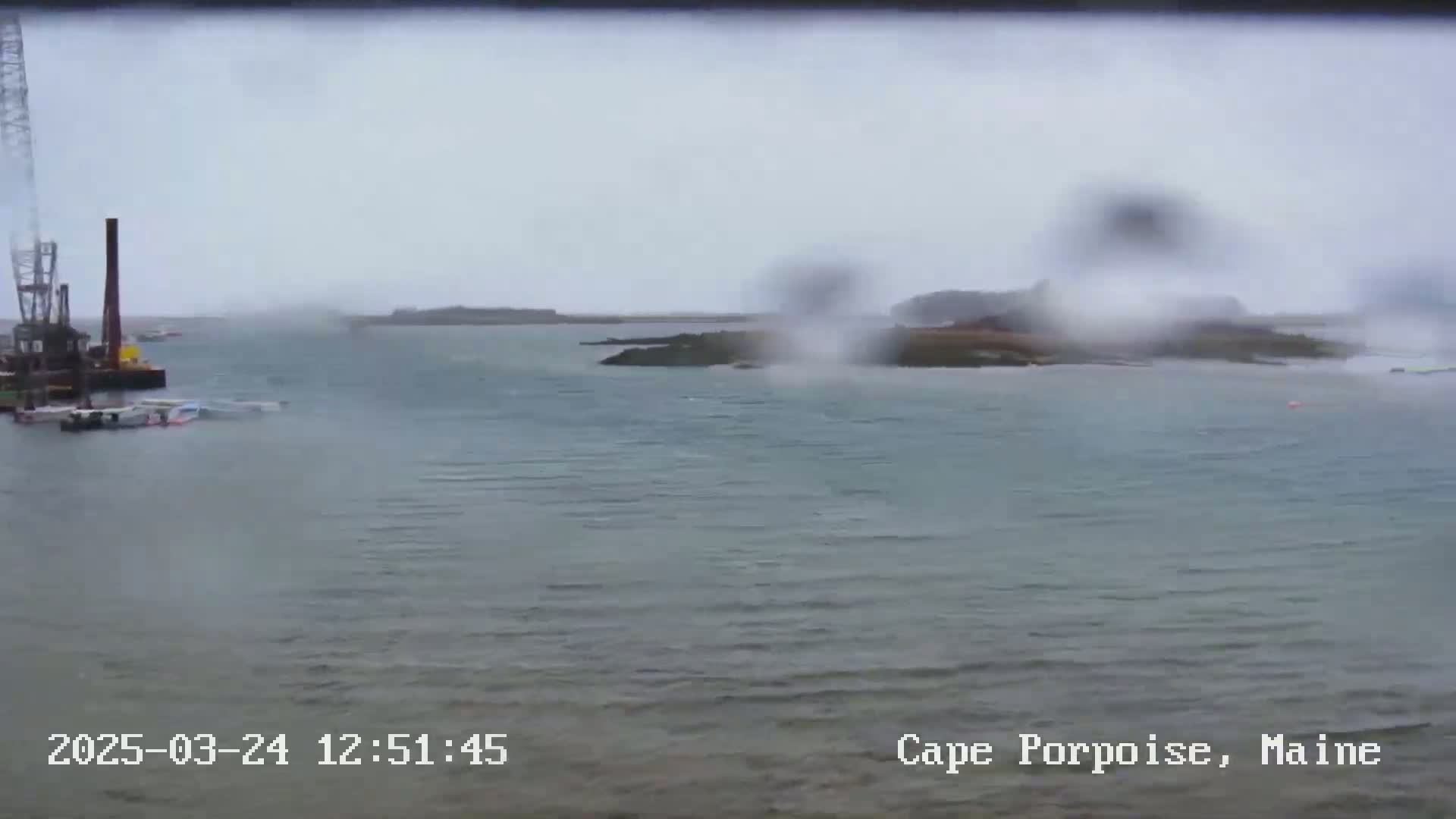 A rainy view of a body of water shows a crane and several boats near the shore, with rocky landmasses visible in the distance.