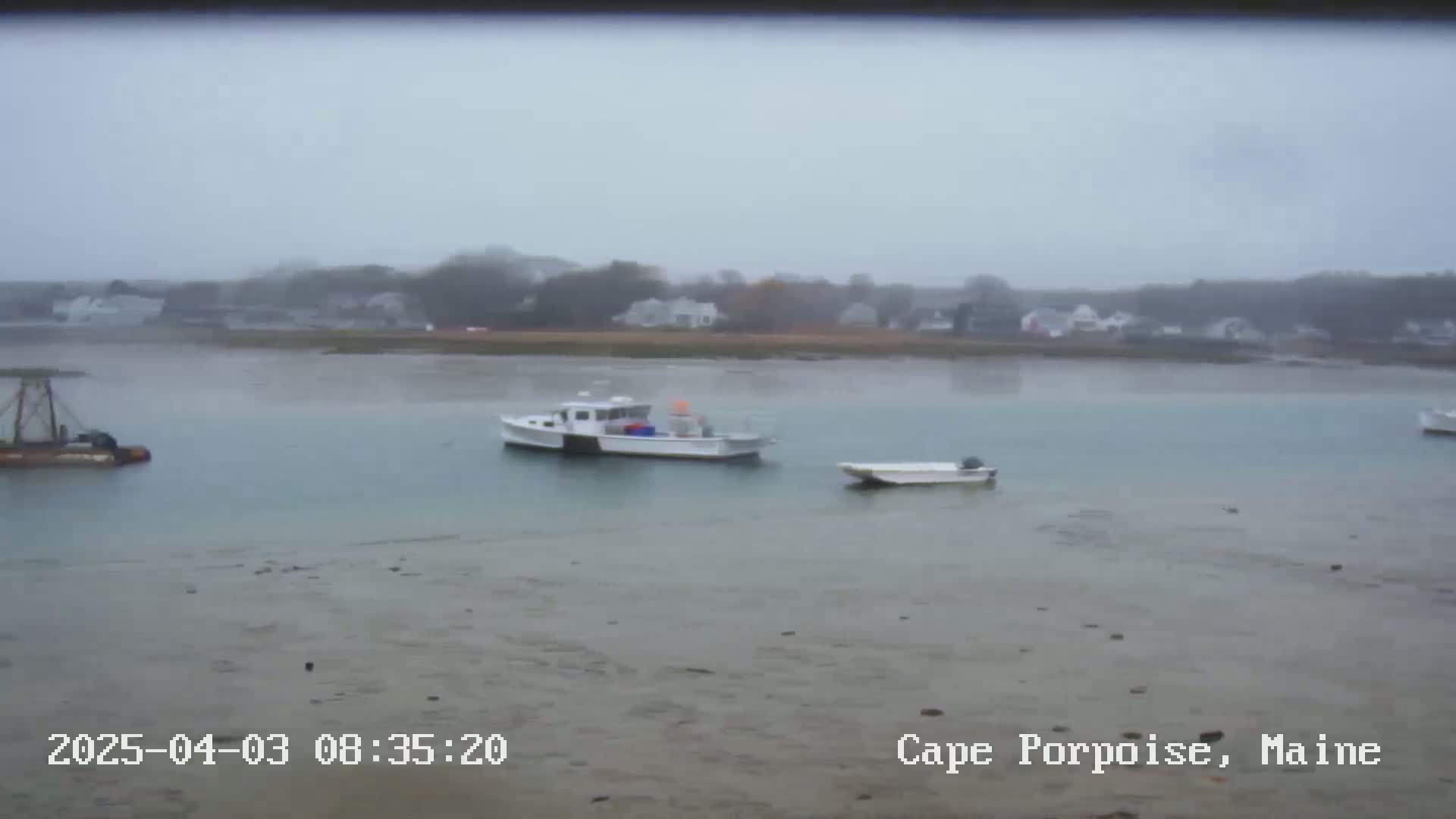 Three boats are moored in a calm, foggy body of water near a shoreline with houses and trees.