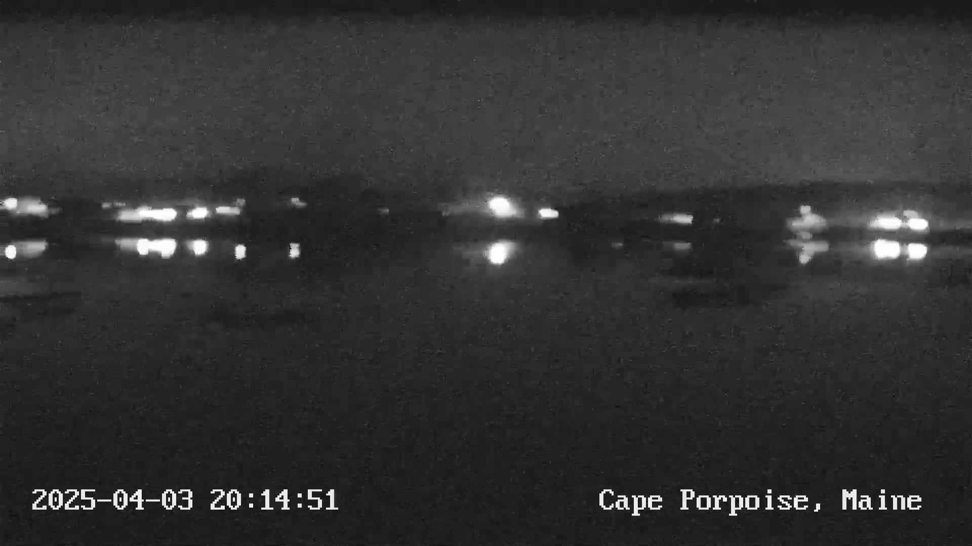 A nighttime view of a dark body of water reflecting the lights of a distant town under a clear sky.