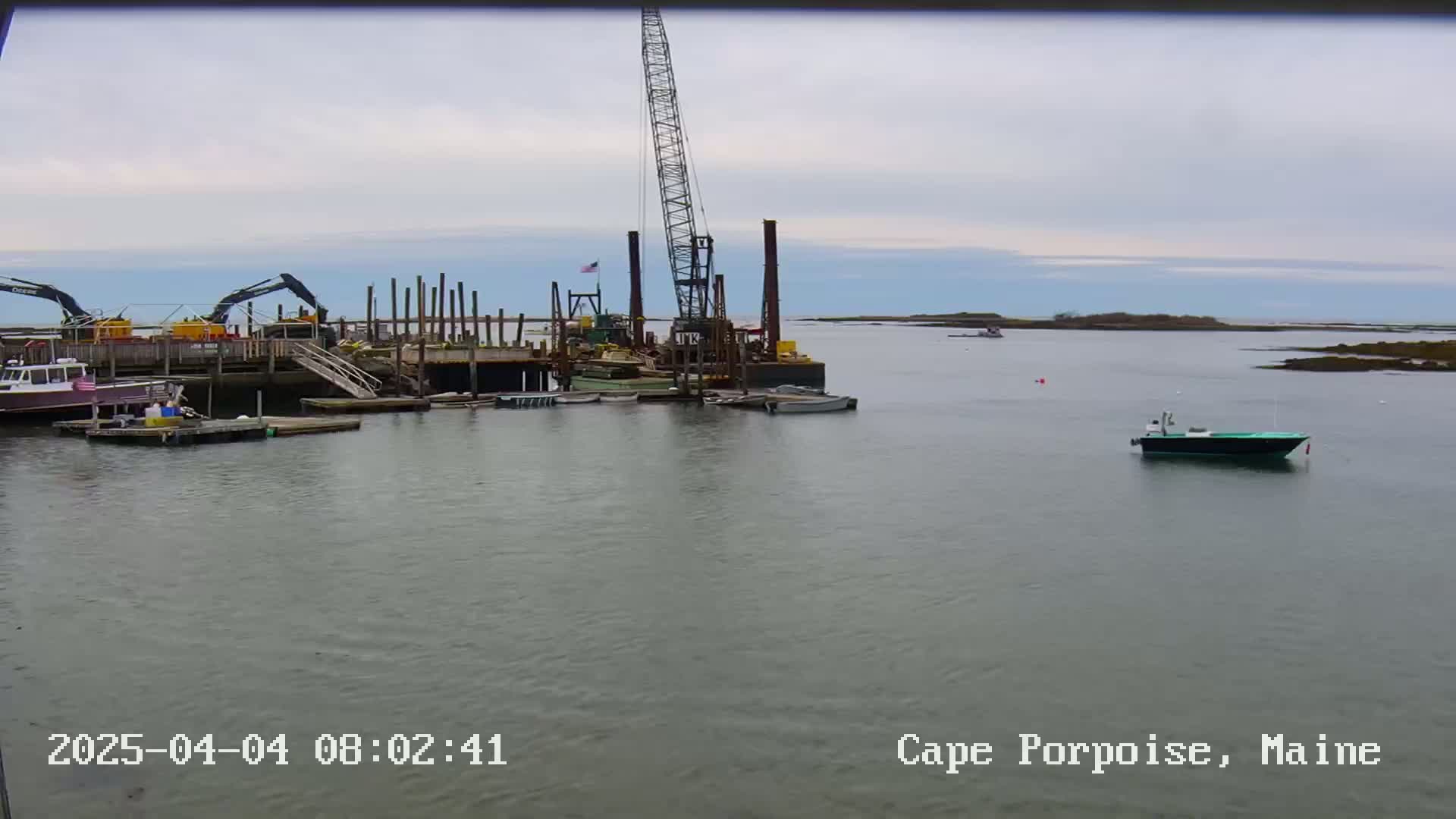 A harbor scene under an overcast sky shows construction activity on a pier with several boats moored nearby.
