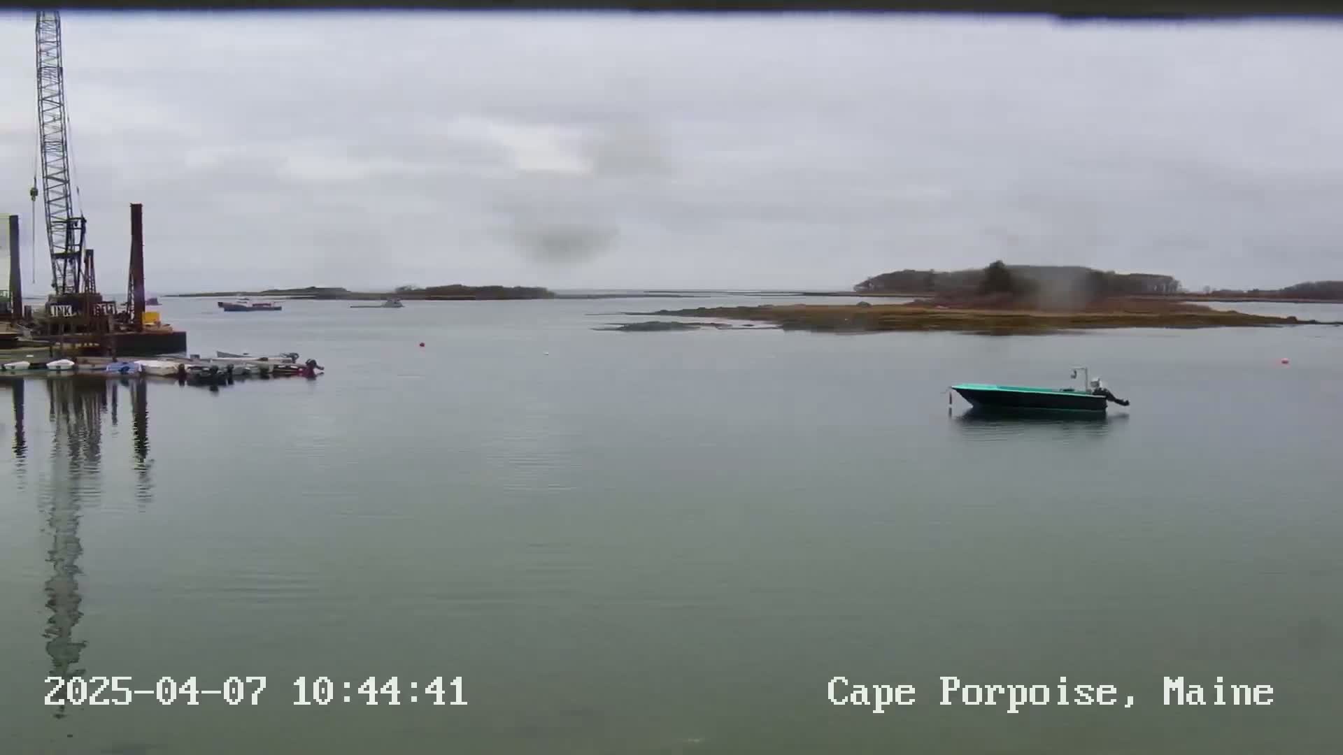 A calm, overcast day shows a body of water with several small boats near a dock and a larger boat further out, with land visible in the distance.