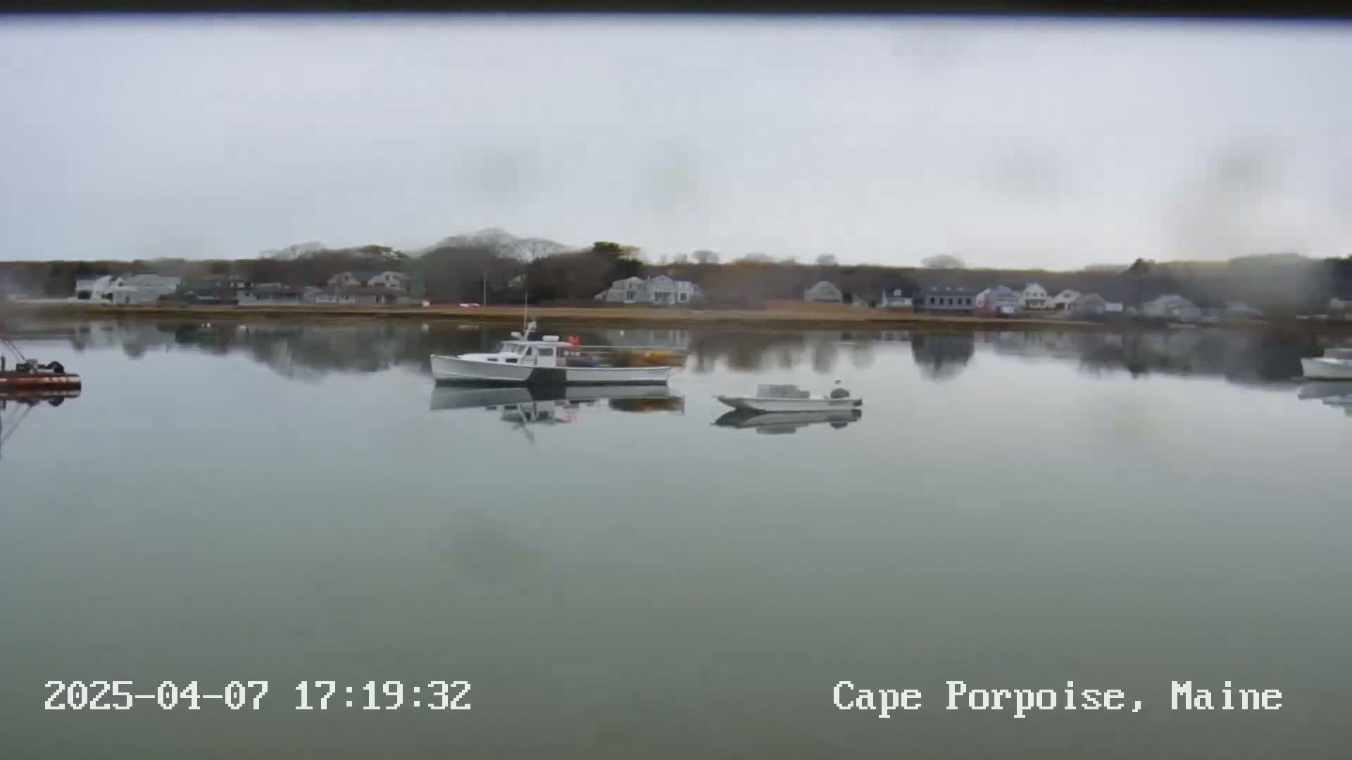 On an overcast day, a calm body of water reflects a shoreline of houses and trees, with several small boats moored in the water.