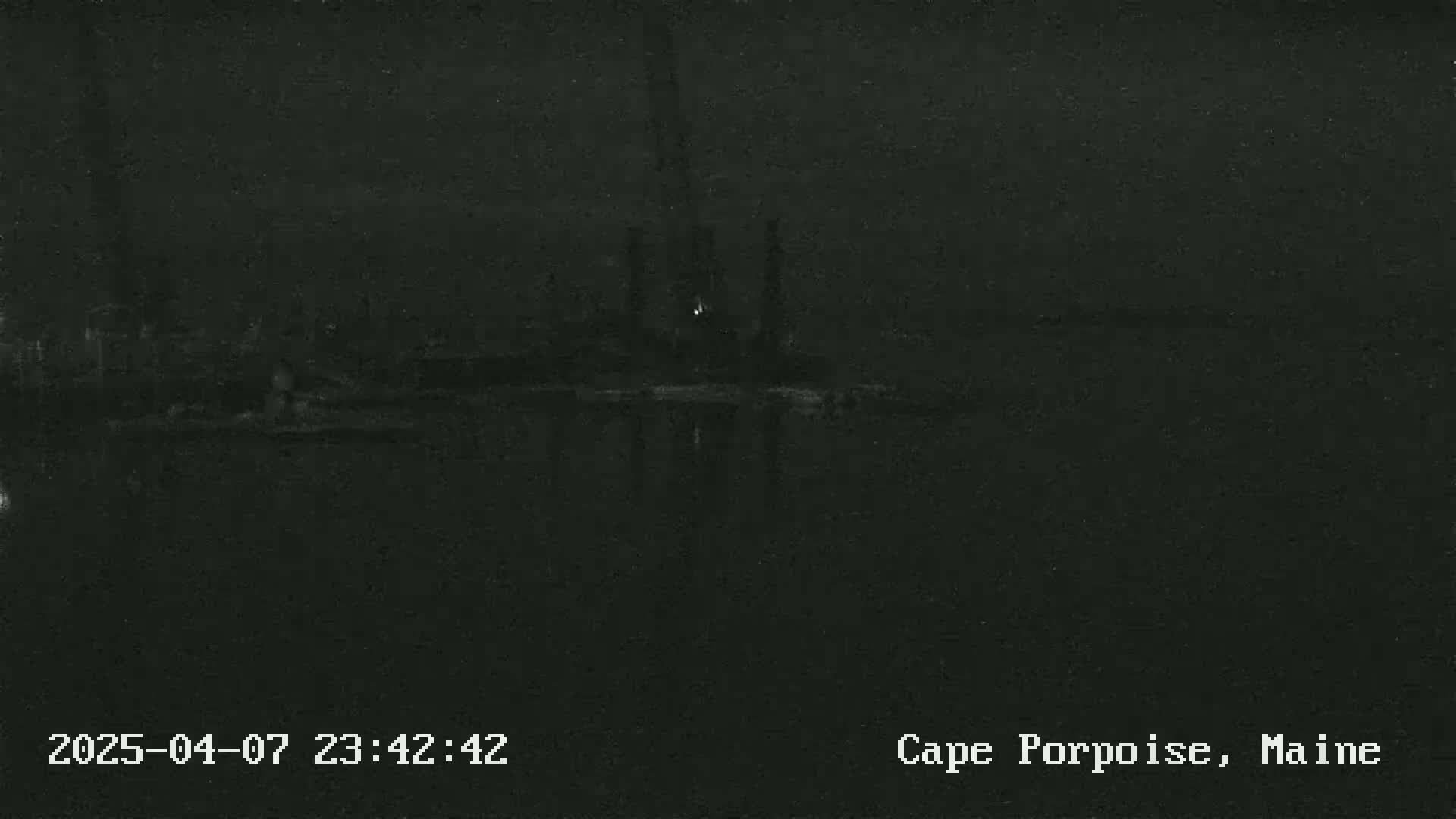 A dark, nighttime view of a waterfront industrial area with faintly visible structures and lights reflected on the water.