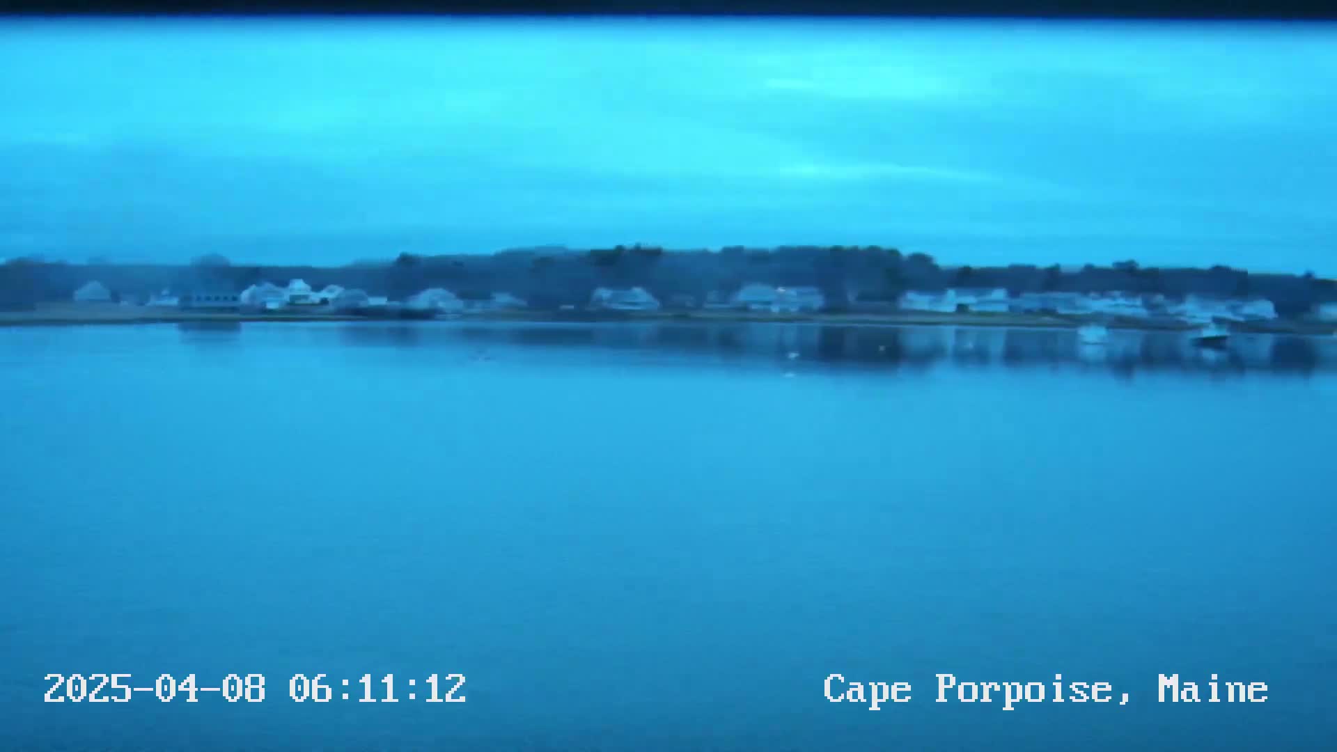 A calm, blue body of water reflects a shoreline of houses and trees under a twilight sky.