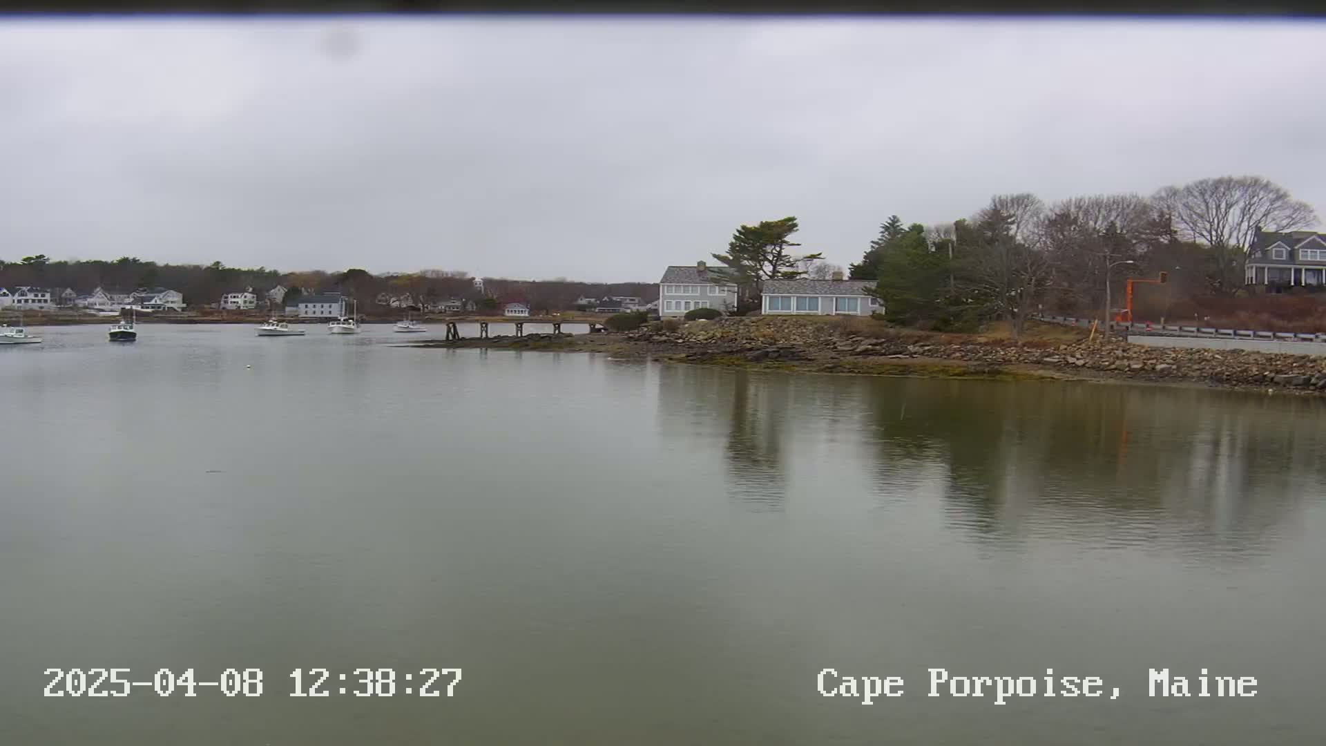 A calm, overcast day shows a harbor with several boats and houses on a rocky shoreline.