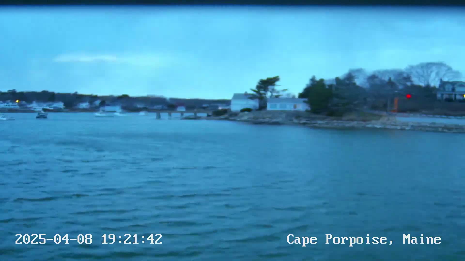 A calm, blue body of water at dusk shows several small boats and houses on a nearby shore under a clear, dusky sky.