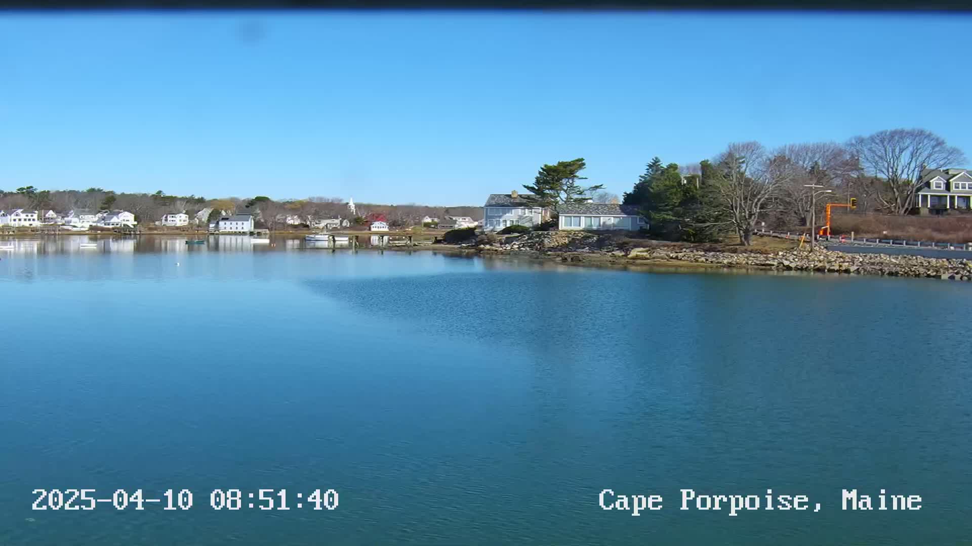 Under a clear blue sky, a calm body of water reflects houses and trees along a shoreline.