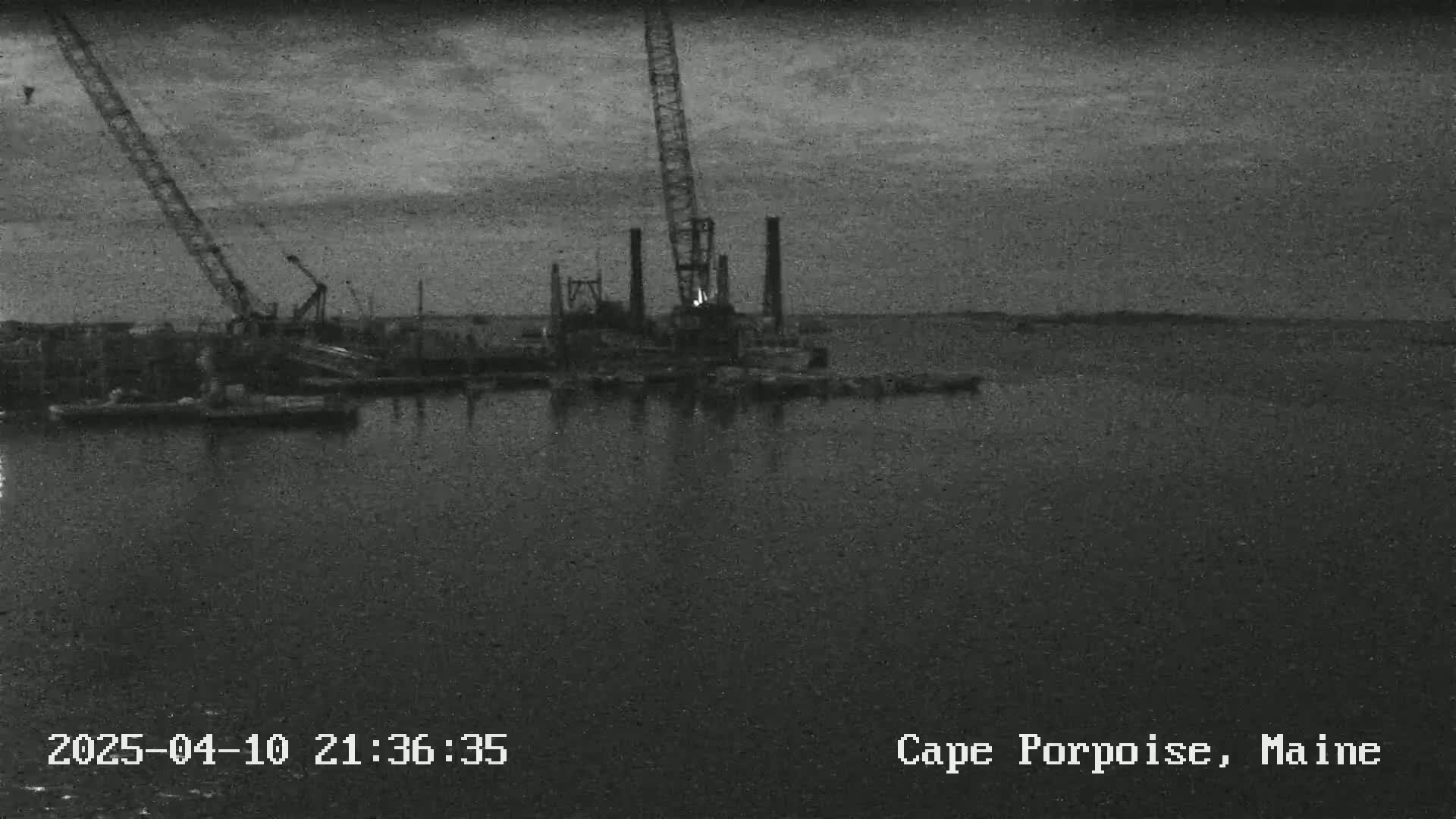 A nighttime, low-light image shows construction equipment and barges on dark water under a cloudy sky.
