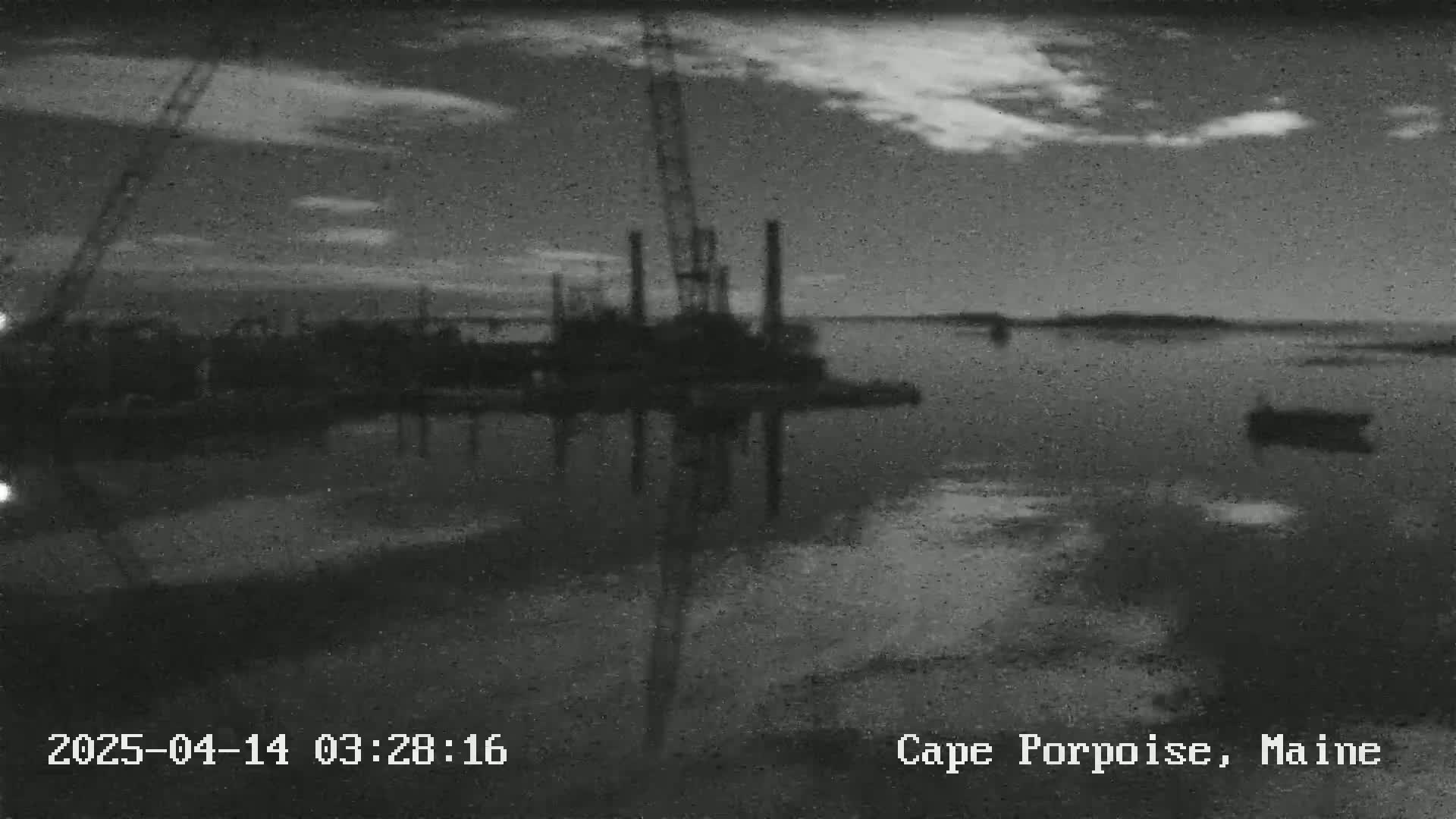 A mostly cloudy sky hangs over a calm body of water reflecting a construction crane and other industrial structures at a waterfront.