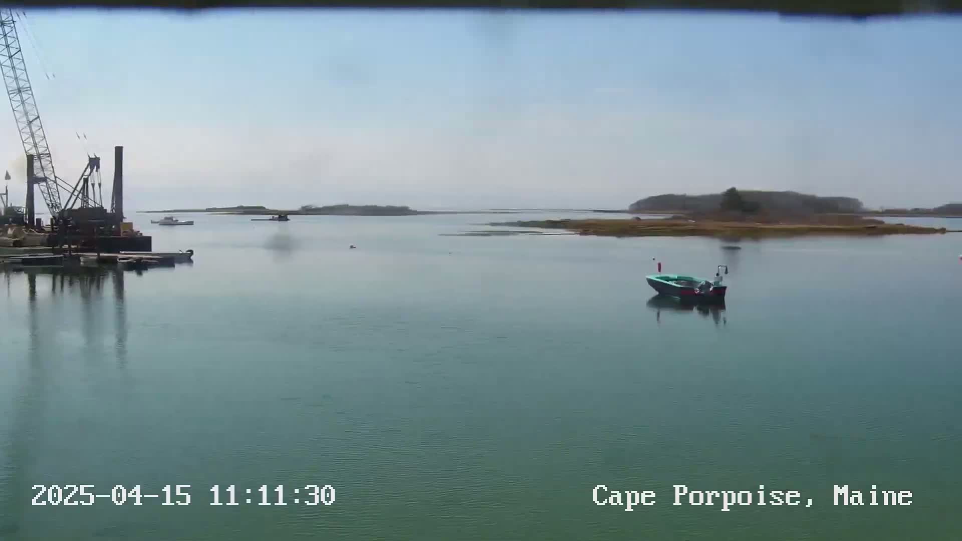 A calm, hazy day shows a body of water with a small boat, a crane and barge in a dock, and land in the background.