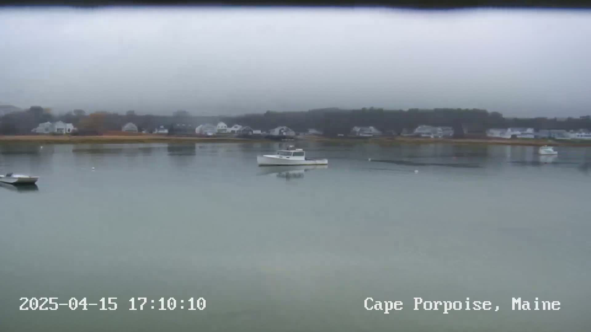 A foggy view of a calm body of water with several small boats and a distant shoreline of houses and trees.