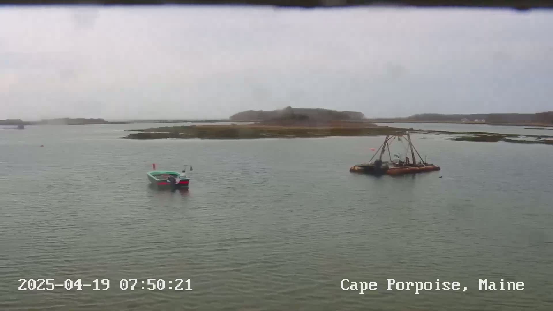 A calm, overcast day shows a small motorboat and a larger dredging vessel in a body of water near a small island.