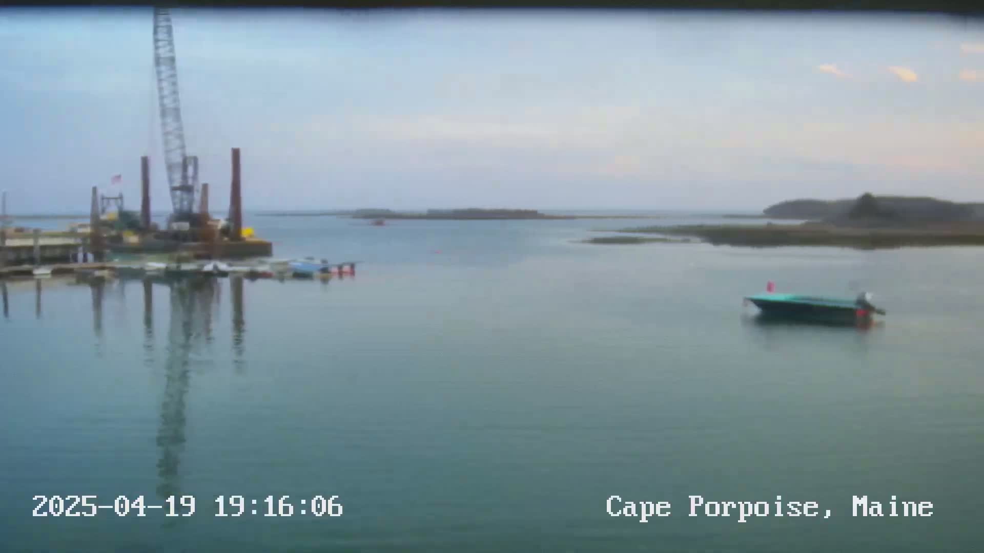 A calm body of water shows a crane and several boats near a dock, with a single boat further out in the water under a mostly cloudy sky.
