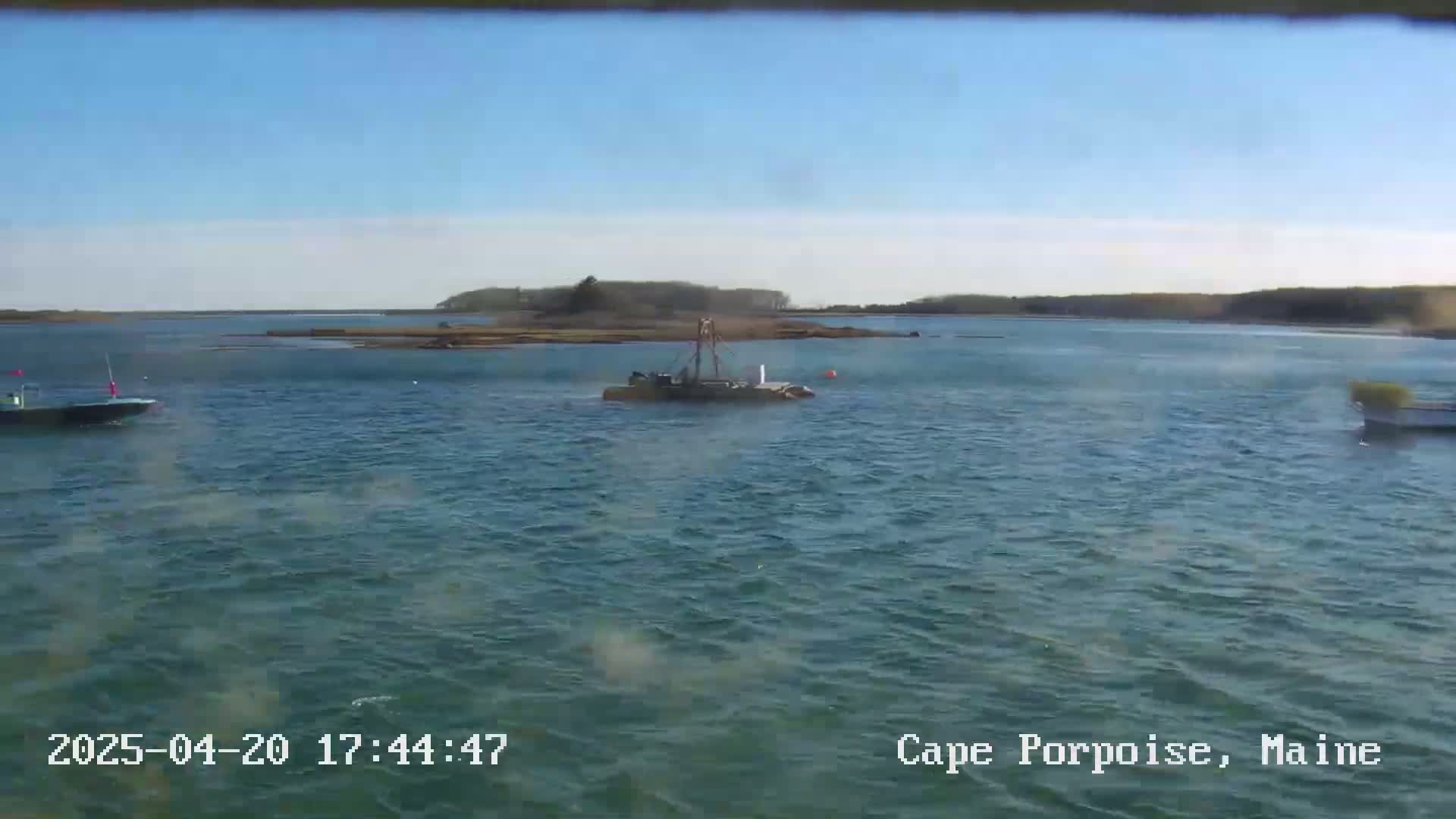 A calm, sunny day shows a body of water with several small boats, one of which appears to be a work boat, and a small island in the background.