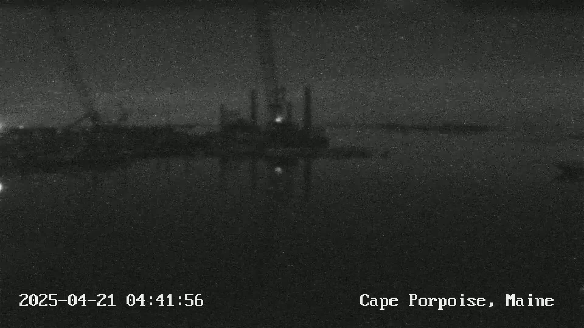 A nighttime, low-light image shows a dark body of water reflecting a dimly lit structure on the far shore.