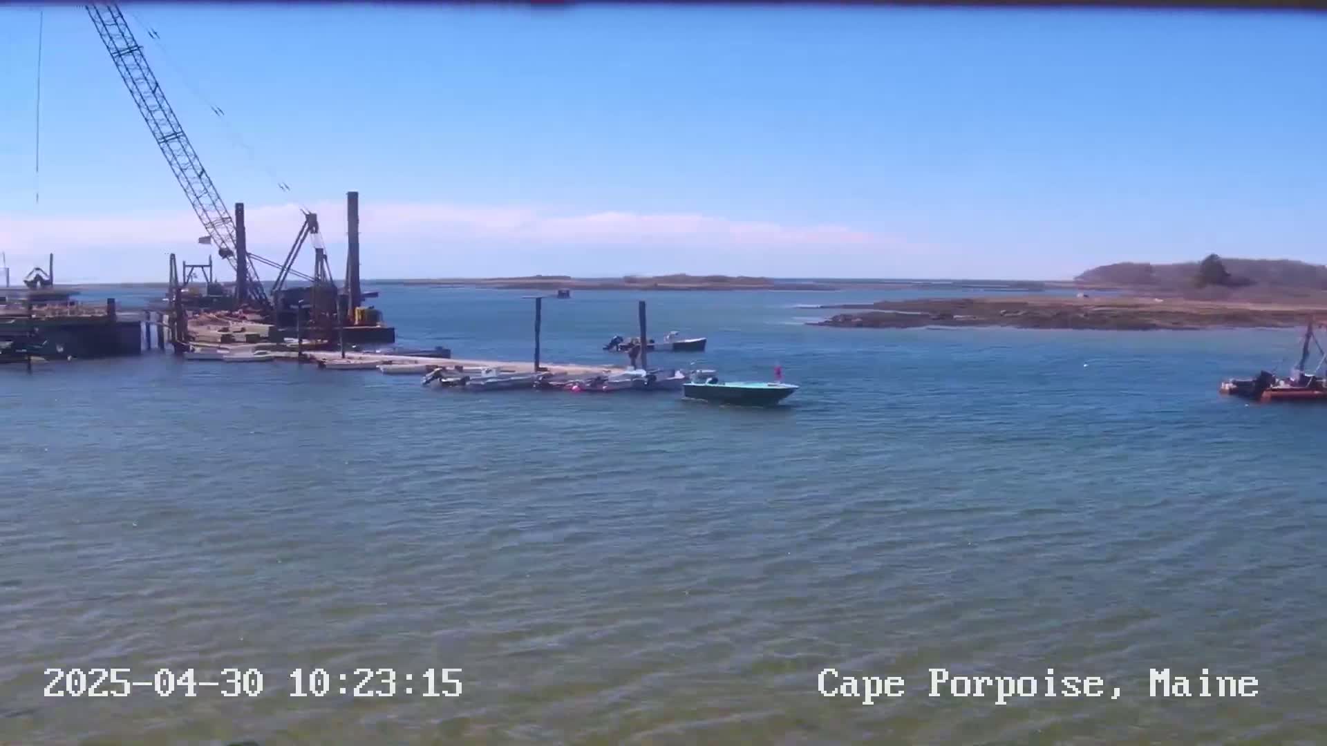 A large crane and several boats are docked at a pier on a calm, sunny day near a rocky shoreline.