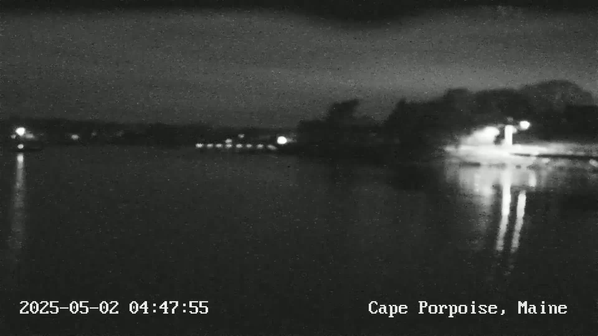 A nighttime view of a dark body of water reflecting lights from the opposite shore under a cloudy sky.