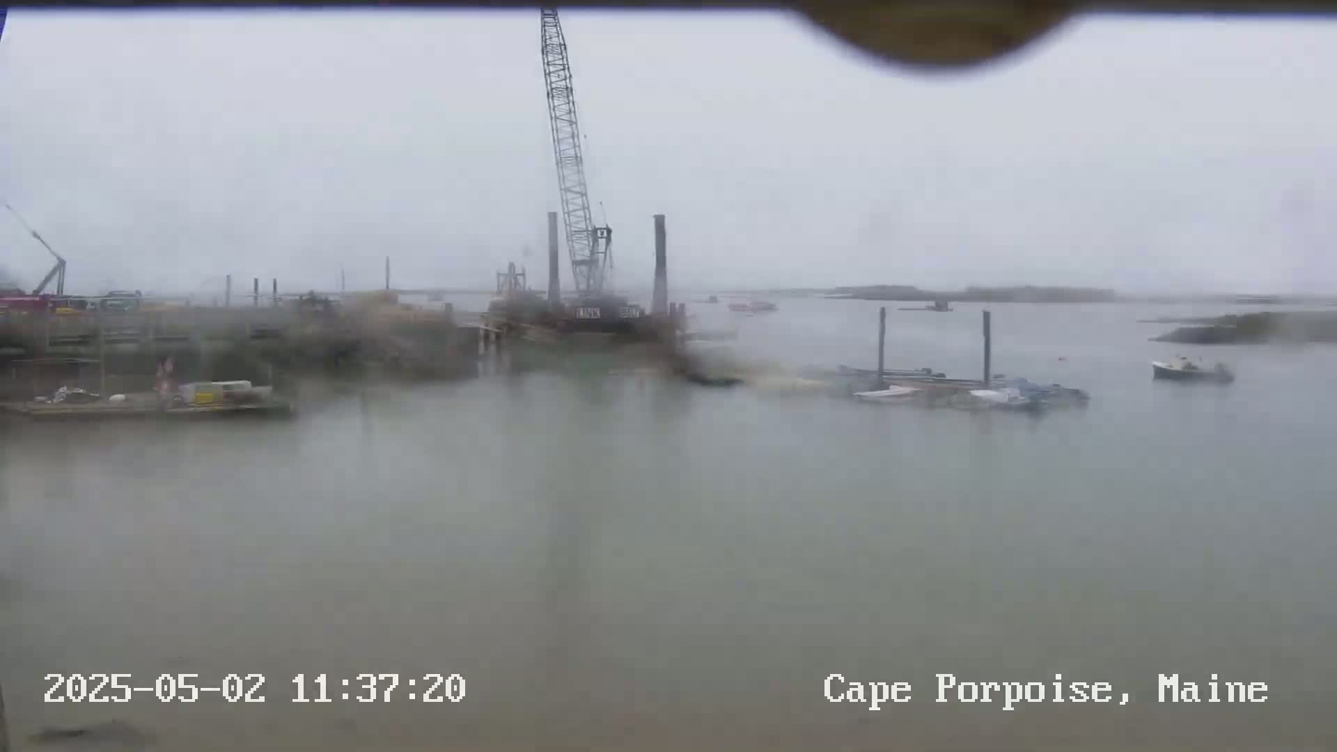 A construction crane and several boats are visible in a harbor on an overcast and rainy day.