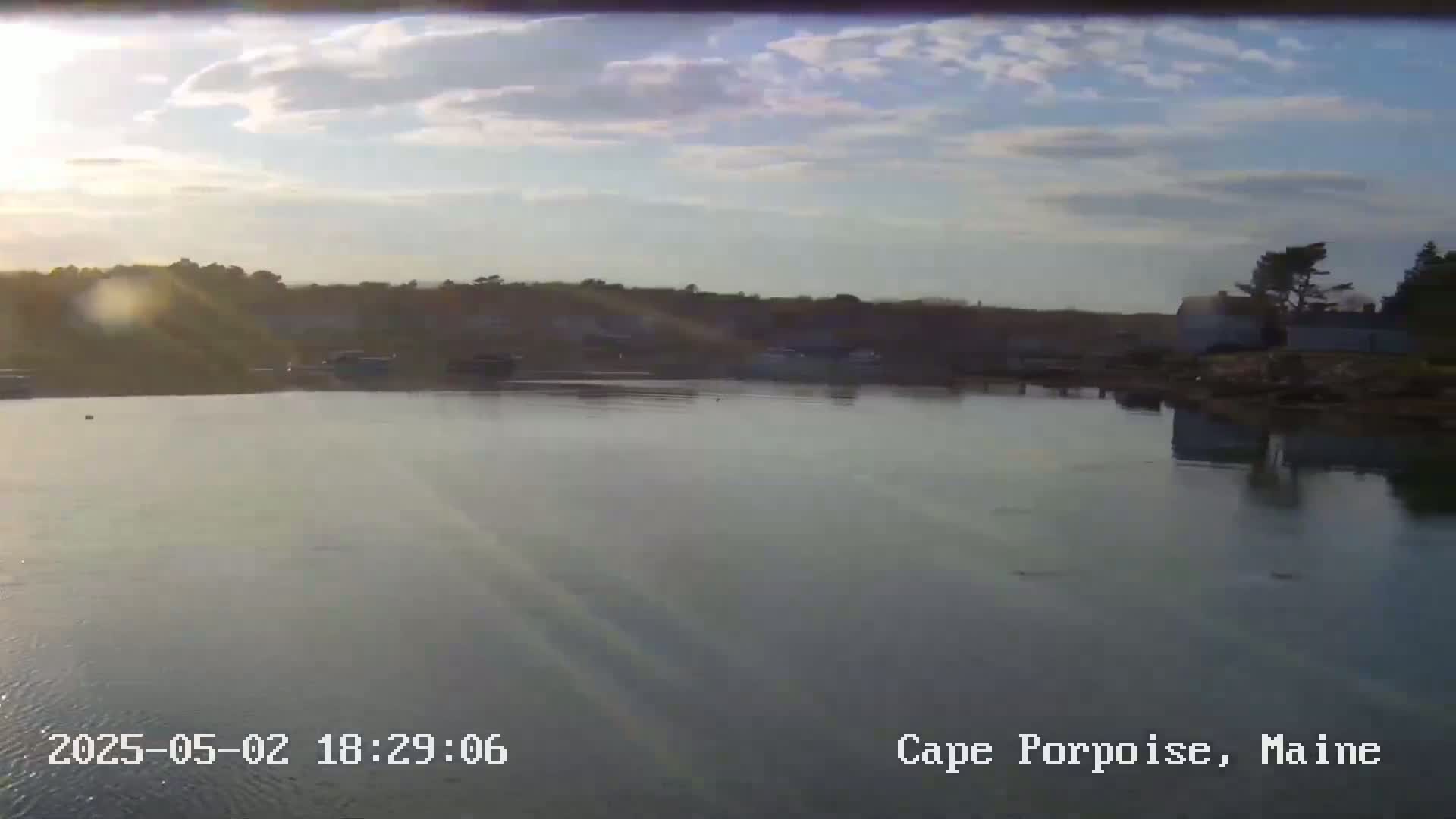 A calm body of water reflects a partly cloudy sunset sky, with a shoreline featuring buildings and trees in the distance.
