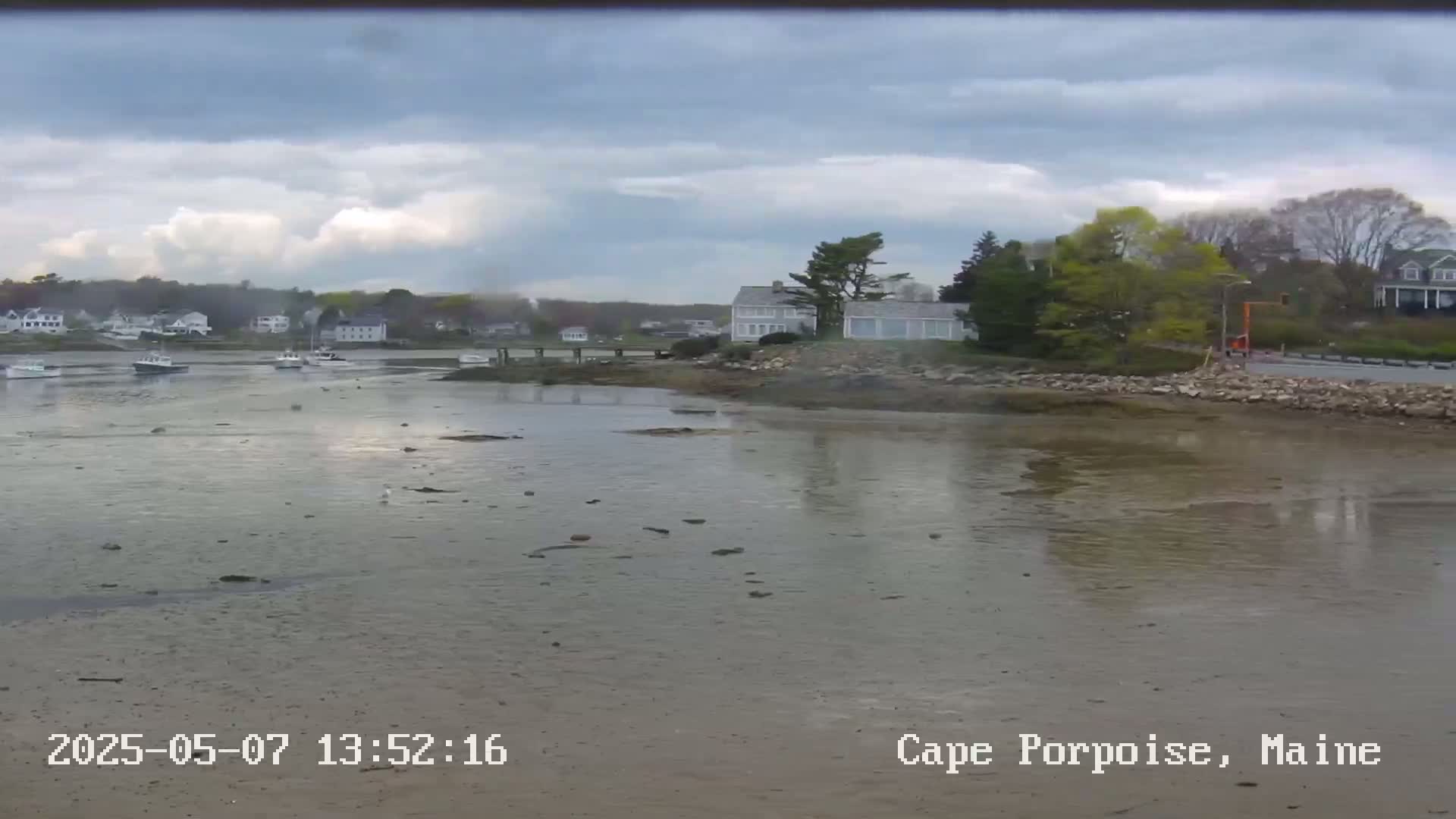 A calm, overcast day reveals a coastal scene with several boats in a harbor, houses on a shoreline, and a mostly exposed mudflat at low tide.