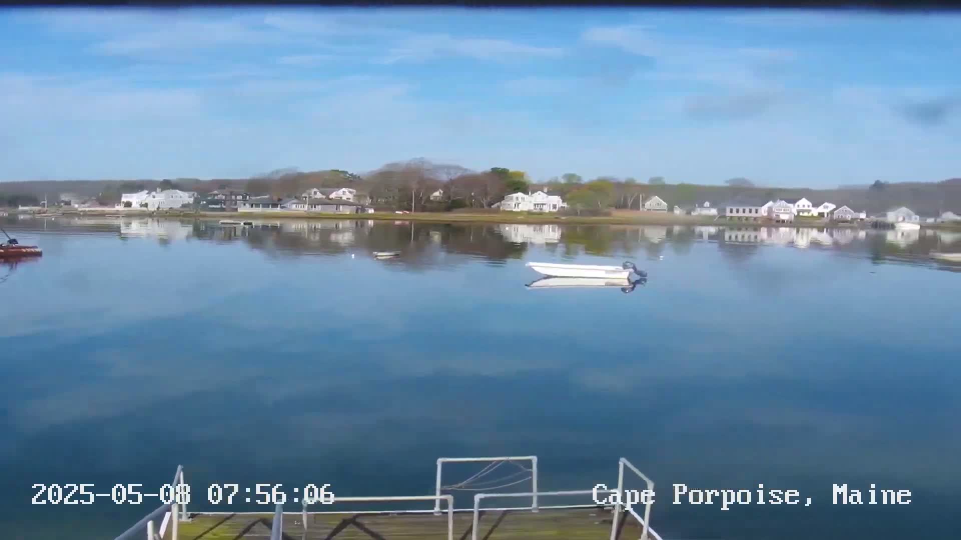 A calm body of water reflects a clear blue sky and houses on the opposite shore, with a small motorboat resting on the water in the foreground.