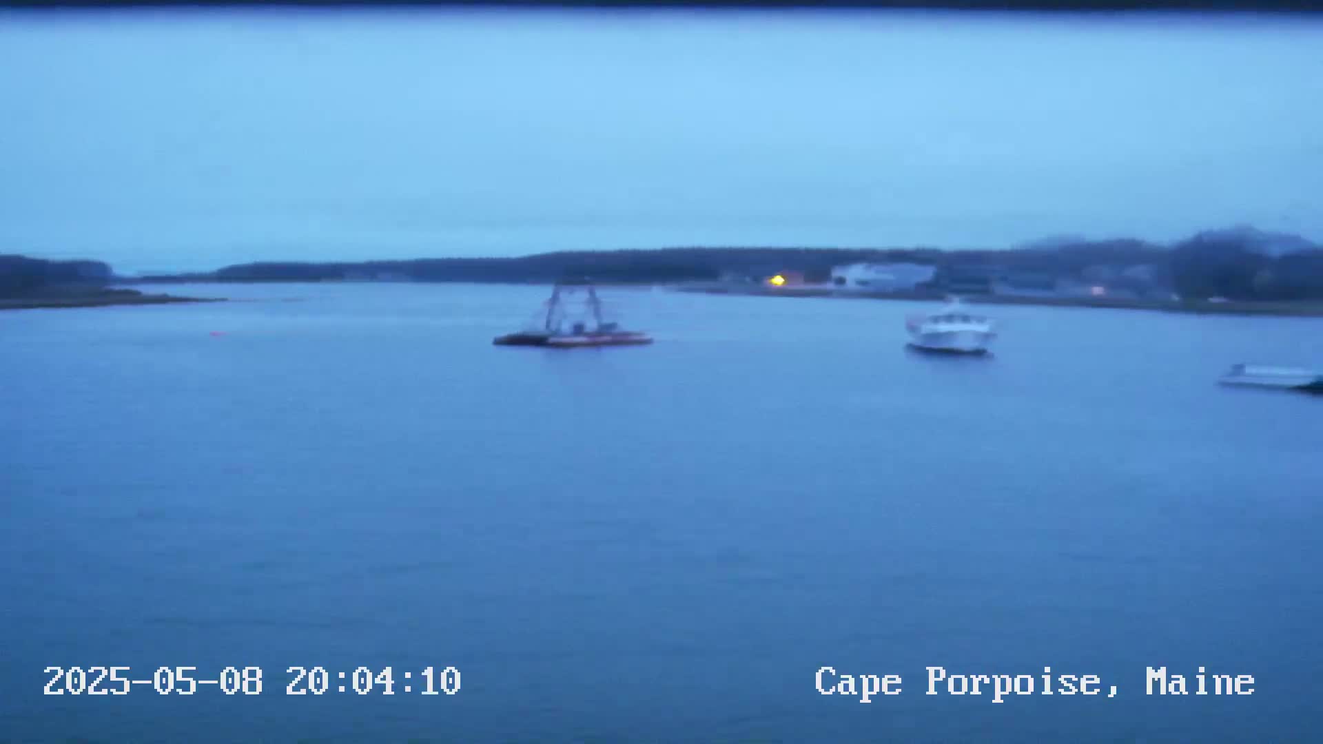 A calm, dusky blue body of water contains several small boats near a dark shoreline under an overcast sky.