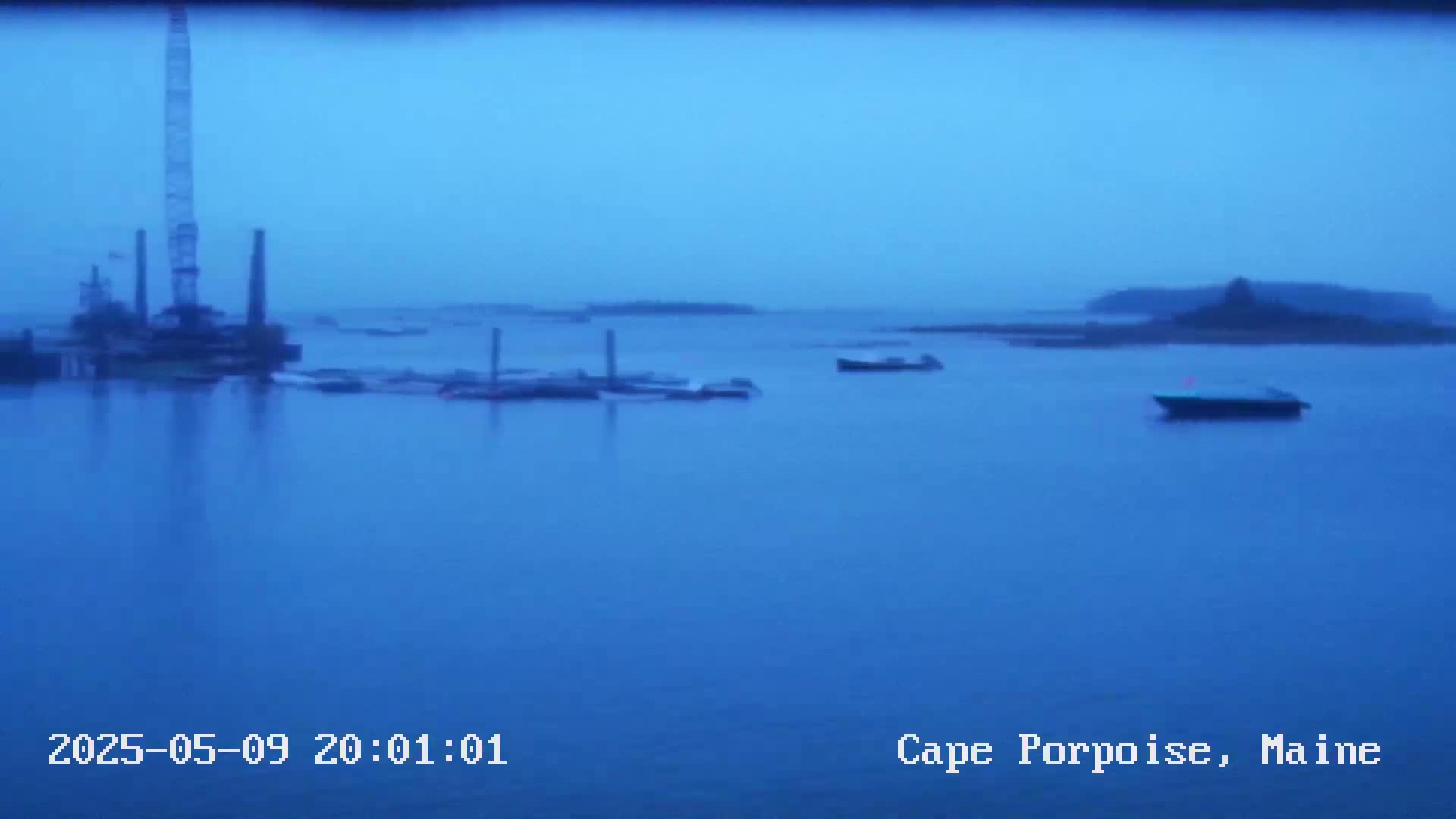 A twilight view of a calm body of water shows several boats and a large industrial structure at a dock, with an island visible in the distance.