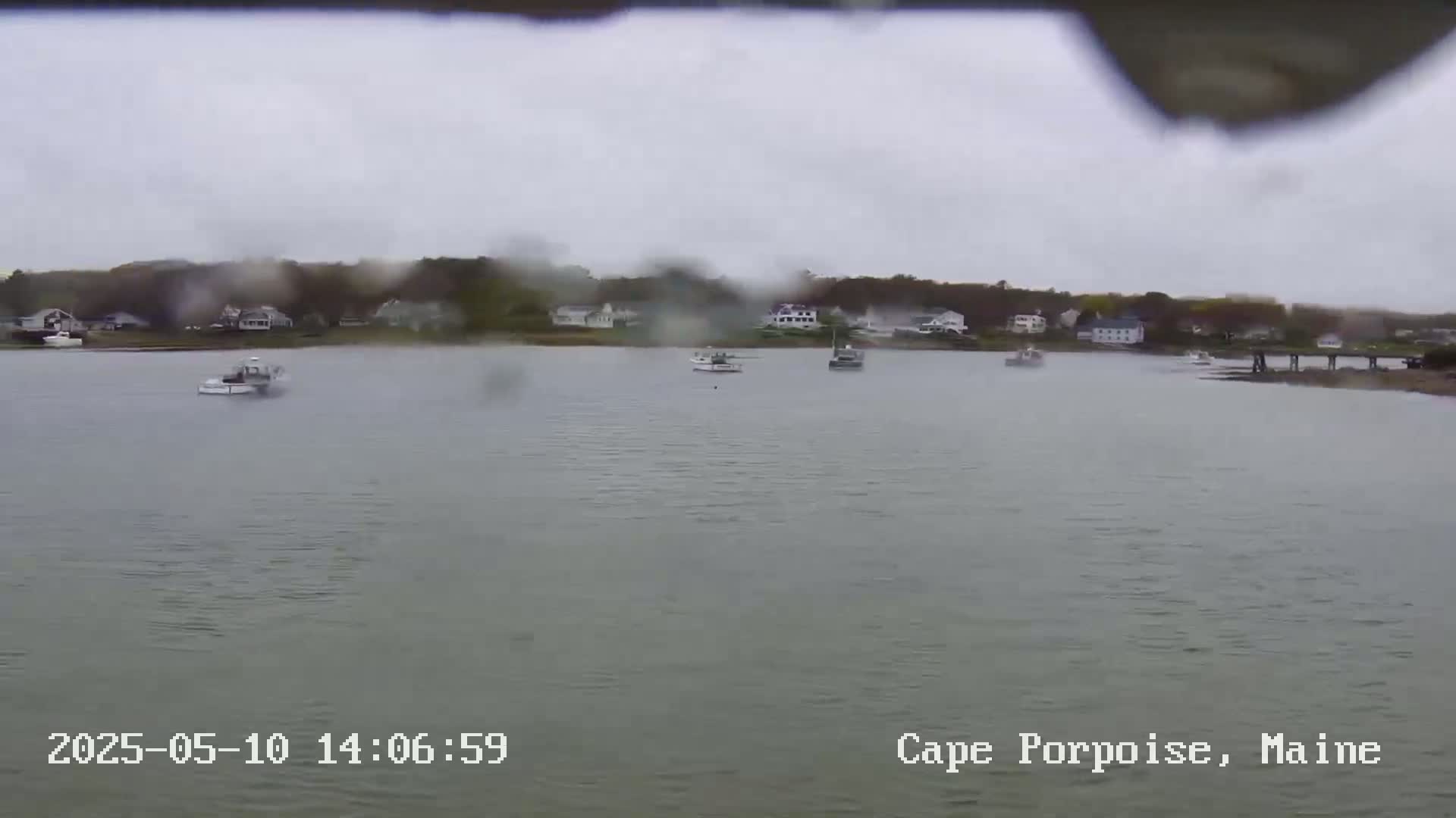 Several boats are moored in a calm body of water in front of a shoreline with houses and trees under an overcast, rainy sky.