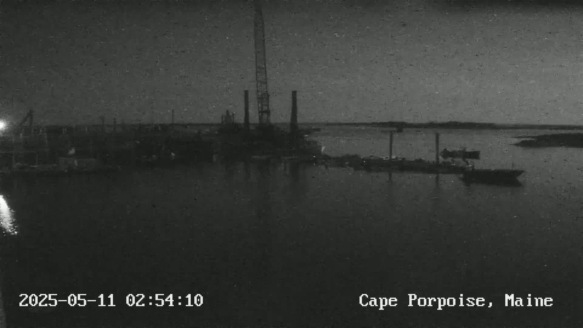 A nighttime, low-light view of a harbor with several boats and a tall crane near industrial structures.