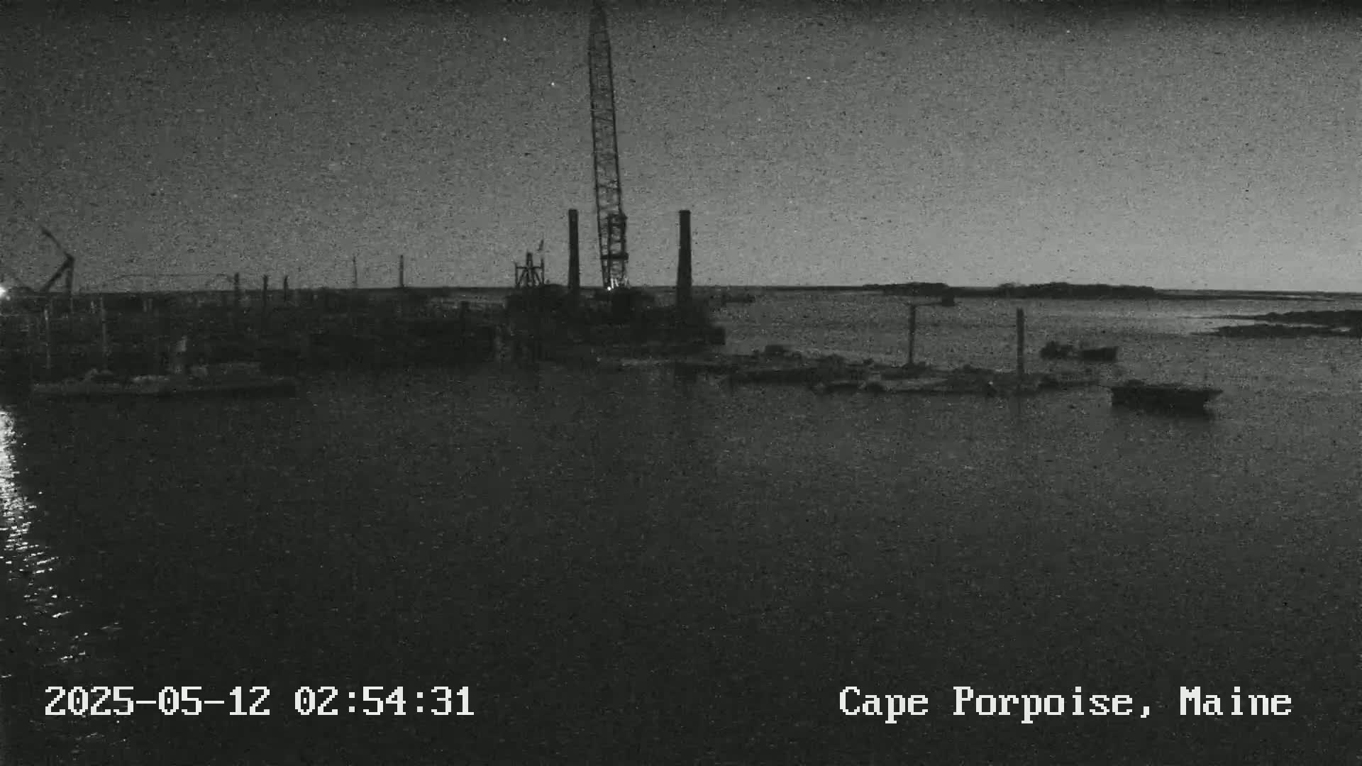 A nighttime, grayscale view shows a harbor with a large crane, several boats, and a calm body of water under a dark sky.