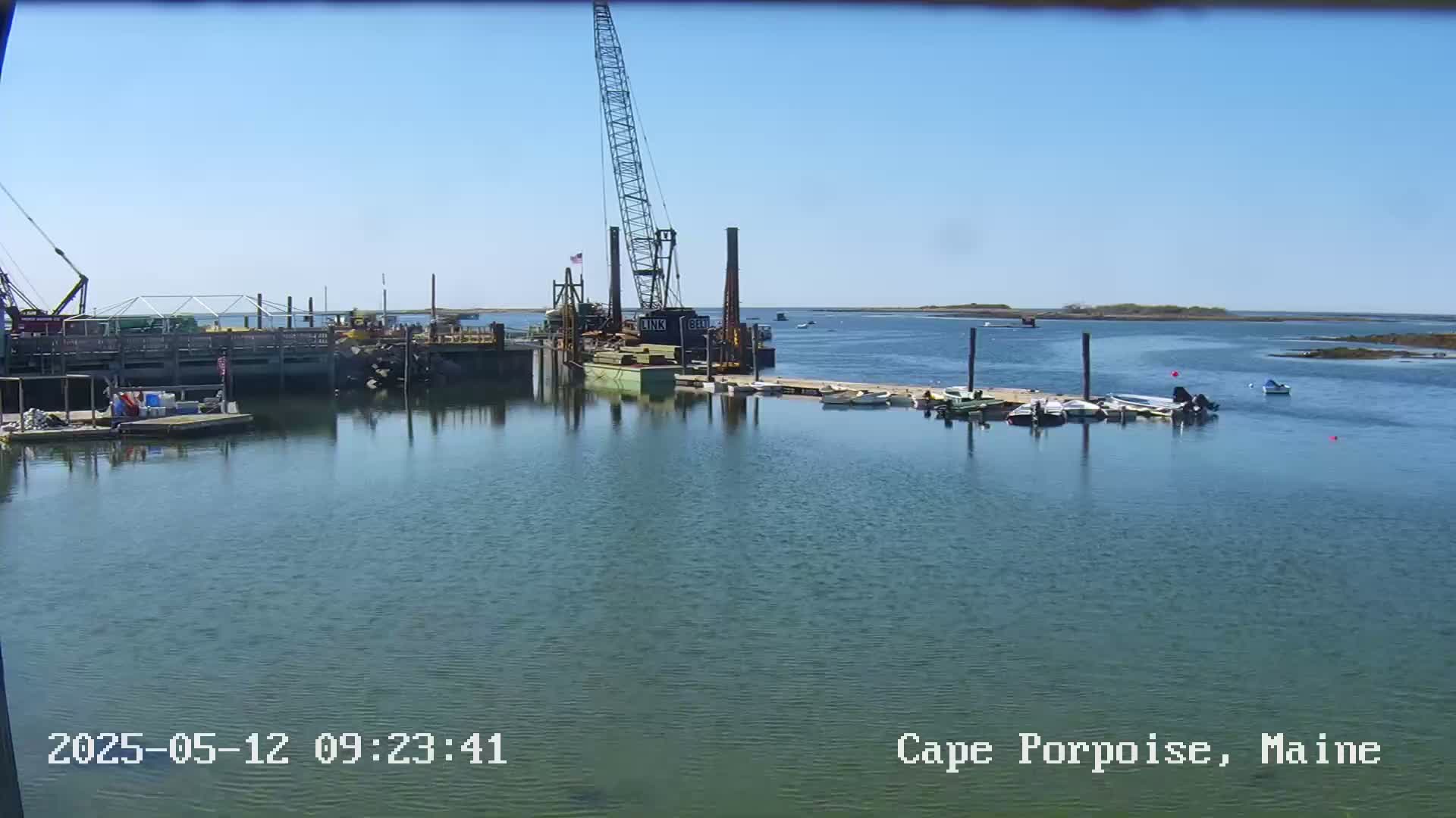 A calm, sunny day shows a harbor with a large crane and barge, several small boats at a dock, and a tranquil body of water extending to a distant shoreline.