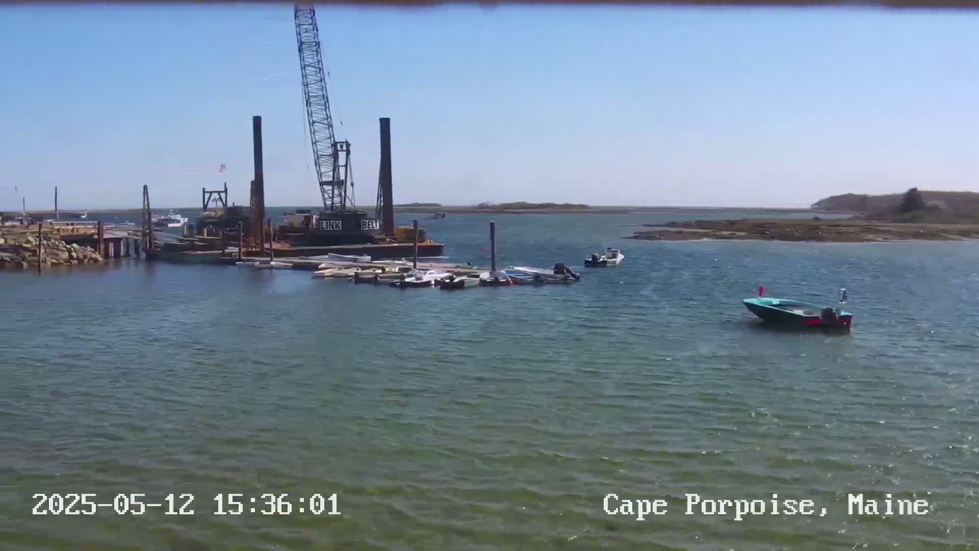 A harbor scene on a sunny day shows a large crane on a barge near several smaller boats moored at a dock, with additional boats in the water beyond.