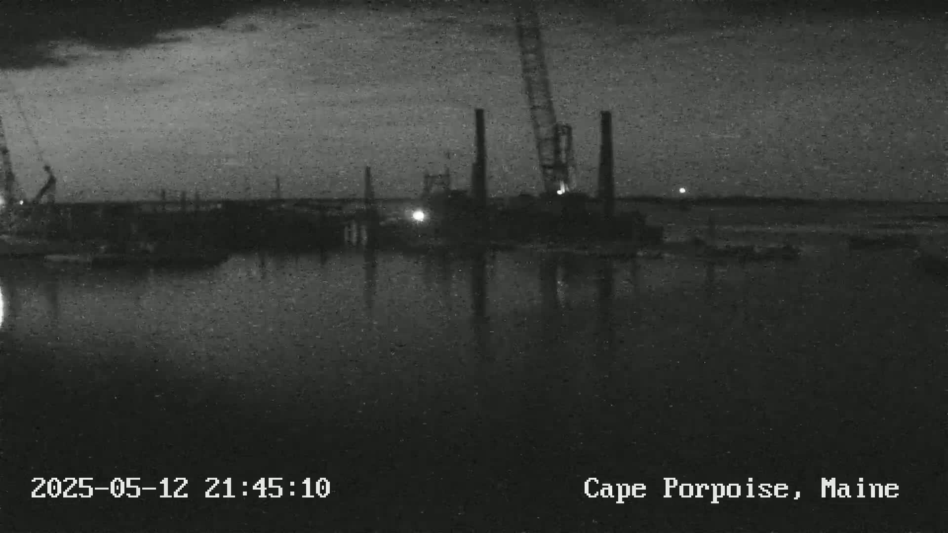 A nighttime, low-light view of a waterfront industrial area with several tall structures and a crane reflected in calm water.