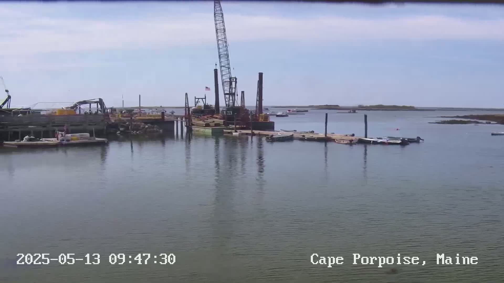 A construction barge with a crane is docked near several other boats on a calm, sunny day.