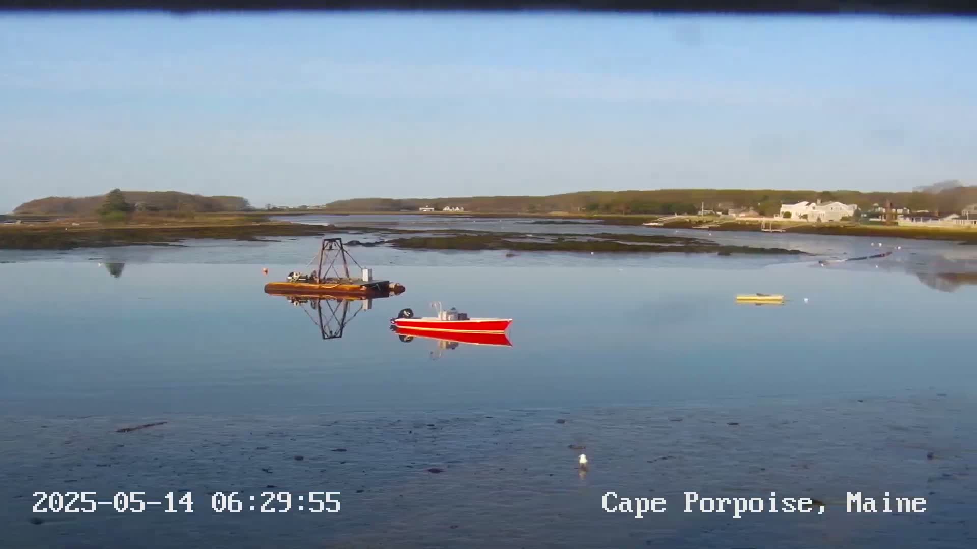 A calm, clear day reveals a tranquil body of water with a small red boat, a barge with a crane, and a yellow boat in the distance, all under a clear blue sky.