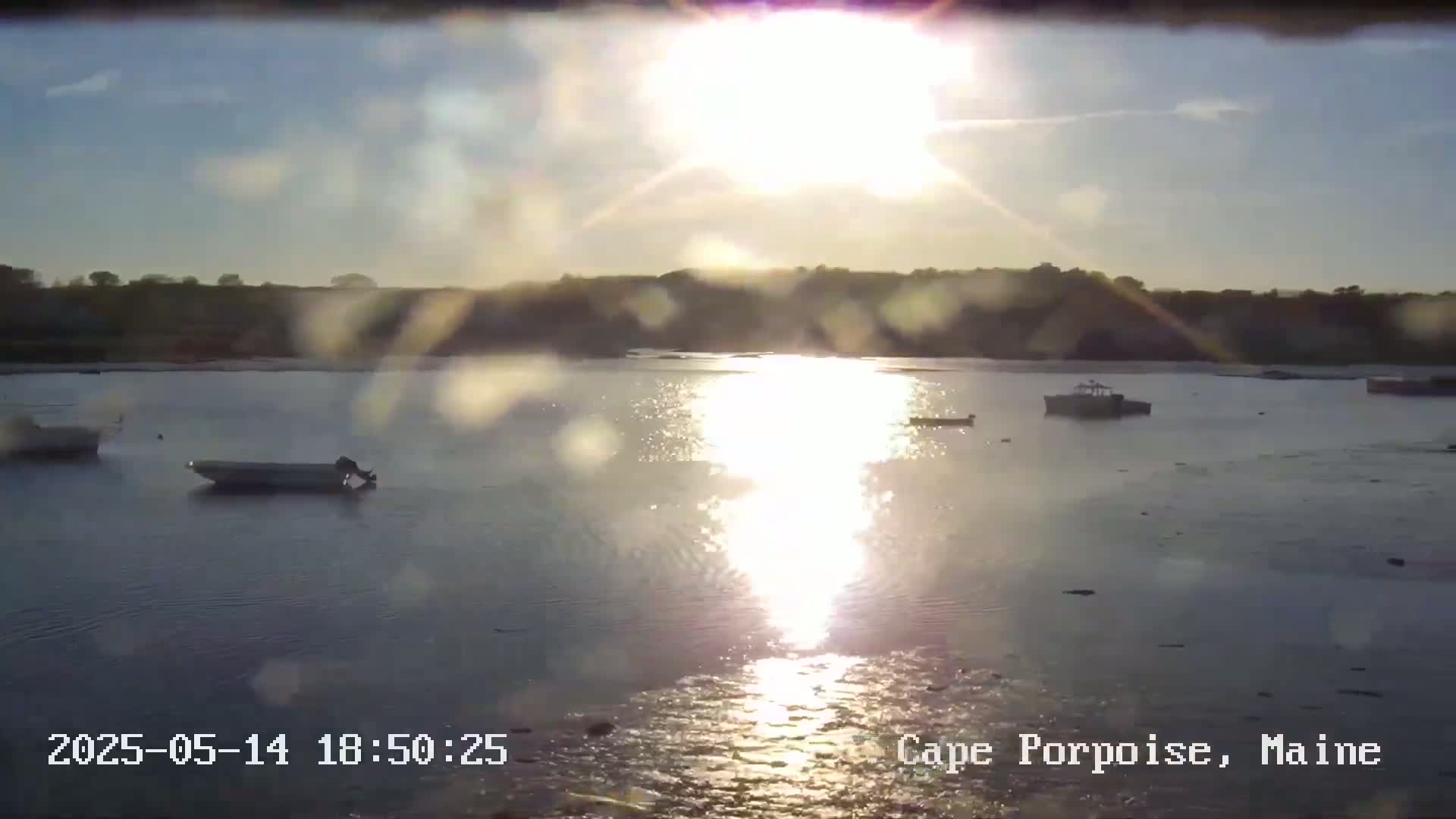 Several small boats are moored in calm water reflecting the bright, hazy sun at sunset, with a distant shoreline visible.
