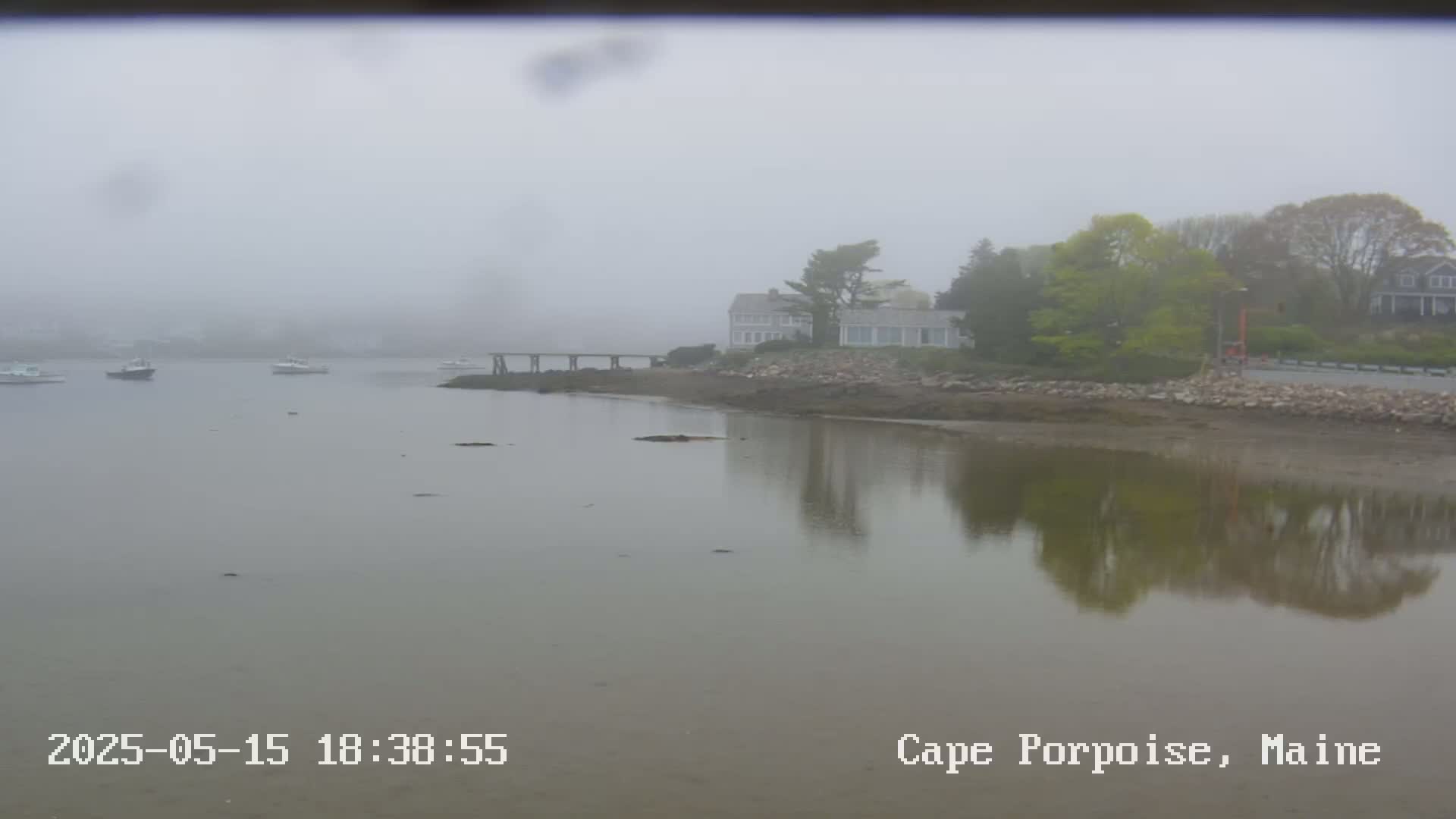A foggy waterfront scene shows several boats in a calm bay, a shoreline with a house and trees, and a pier extending into the water.