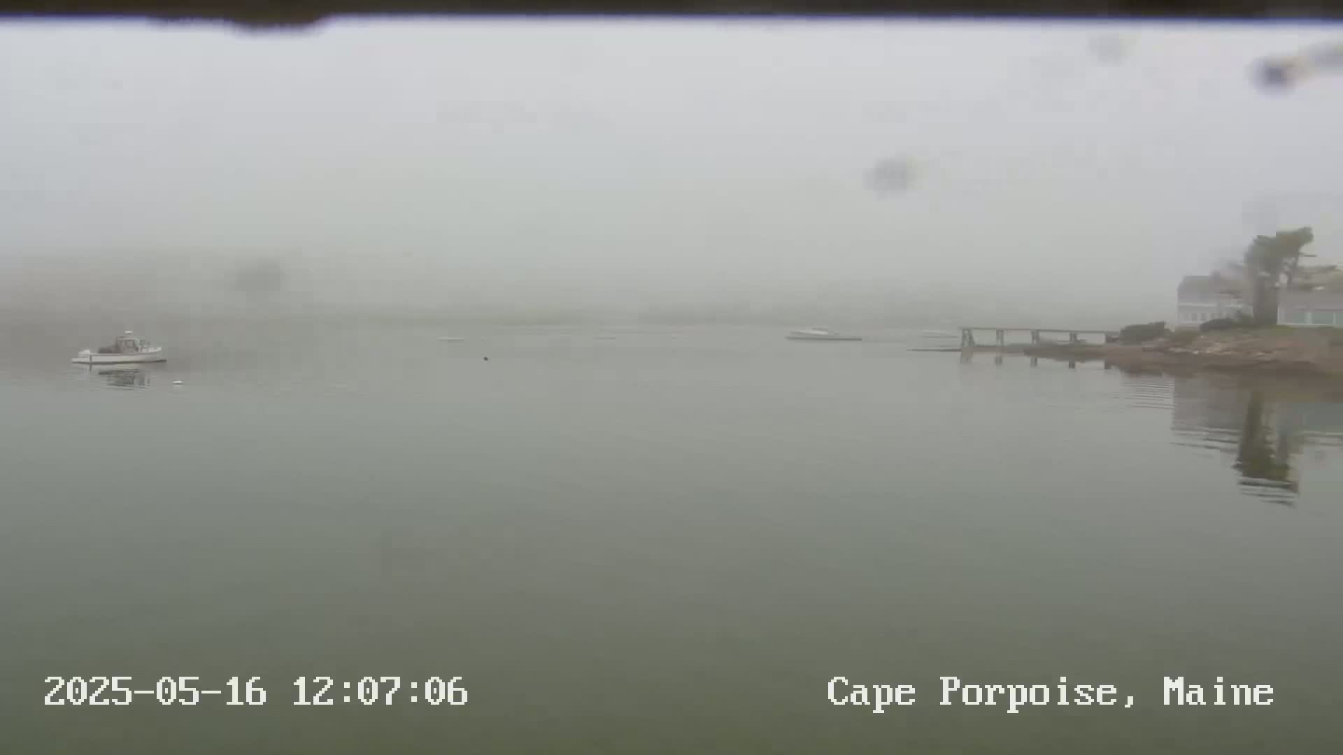 A foggy view of a calm body of water with a small boat and a distant shoreline featuring a pier and houses.