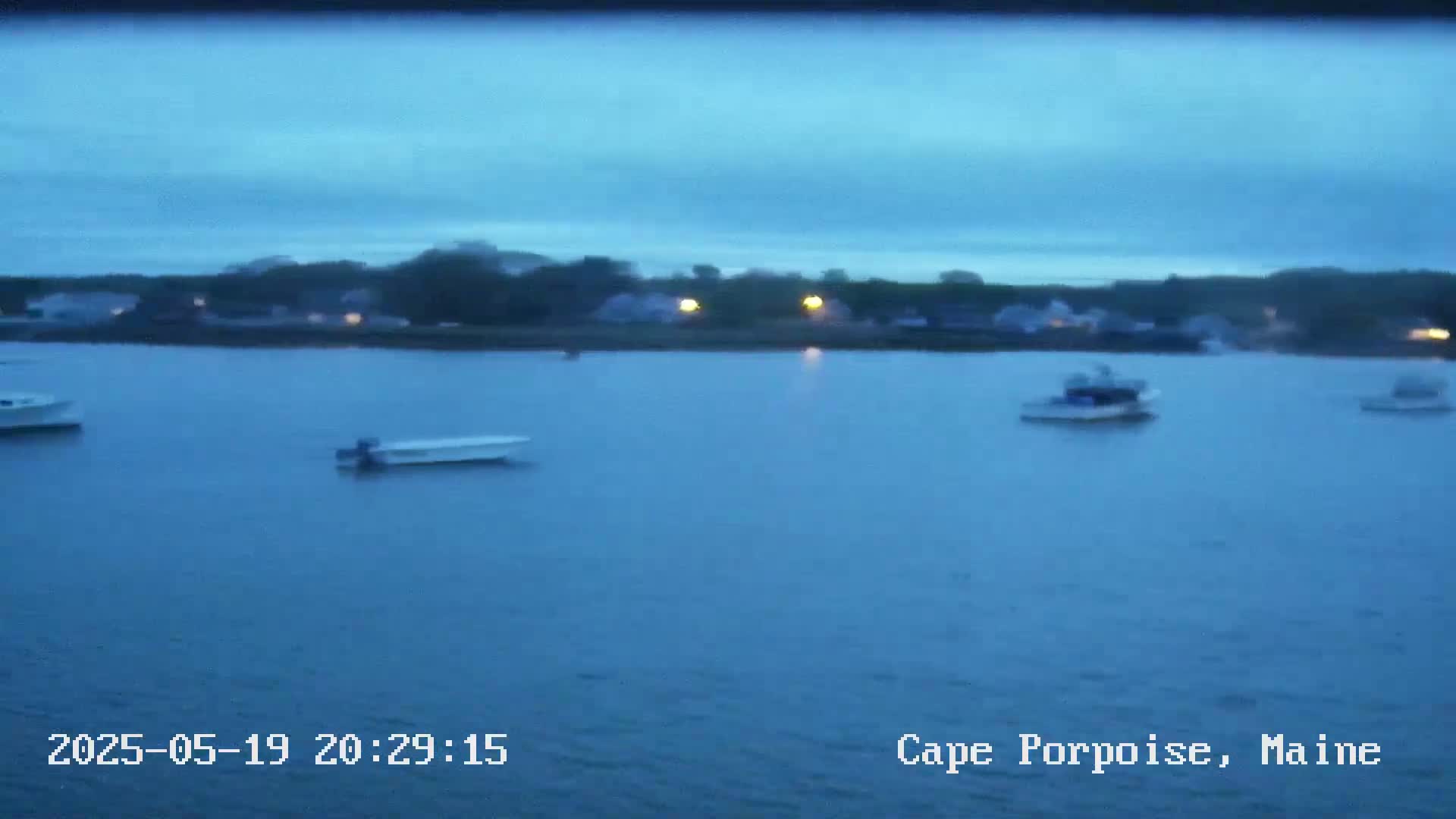 Several small boats are moored in calm, dark blue water in front of a dimly lit shoreline under a twilight sky.