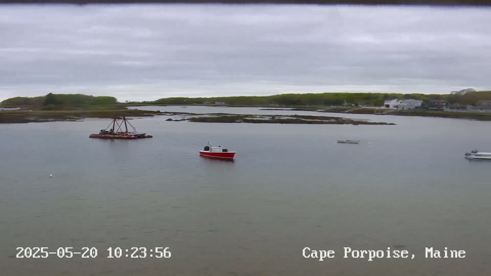 A calm body of water shows a few small boats, a barge with equipment, and a shoreline with green trees under an overcast sky.